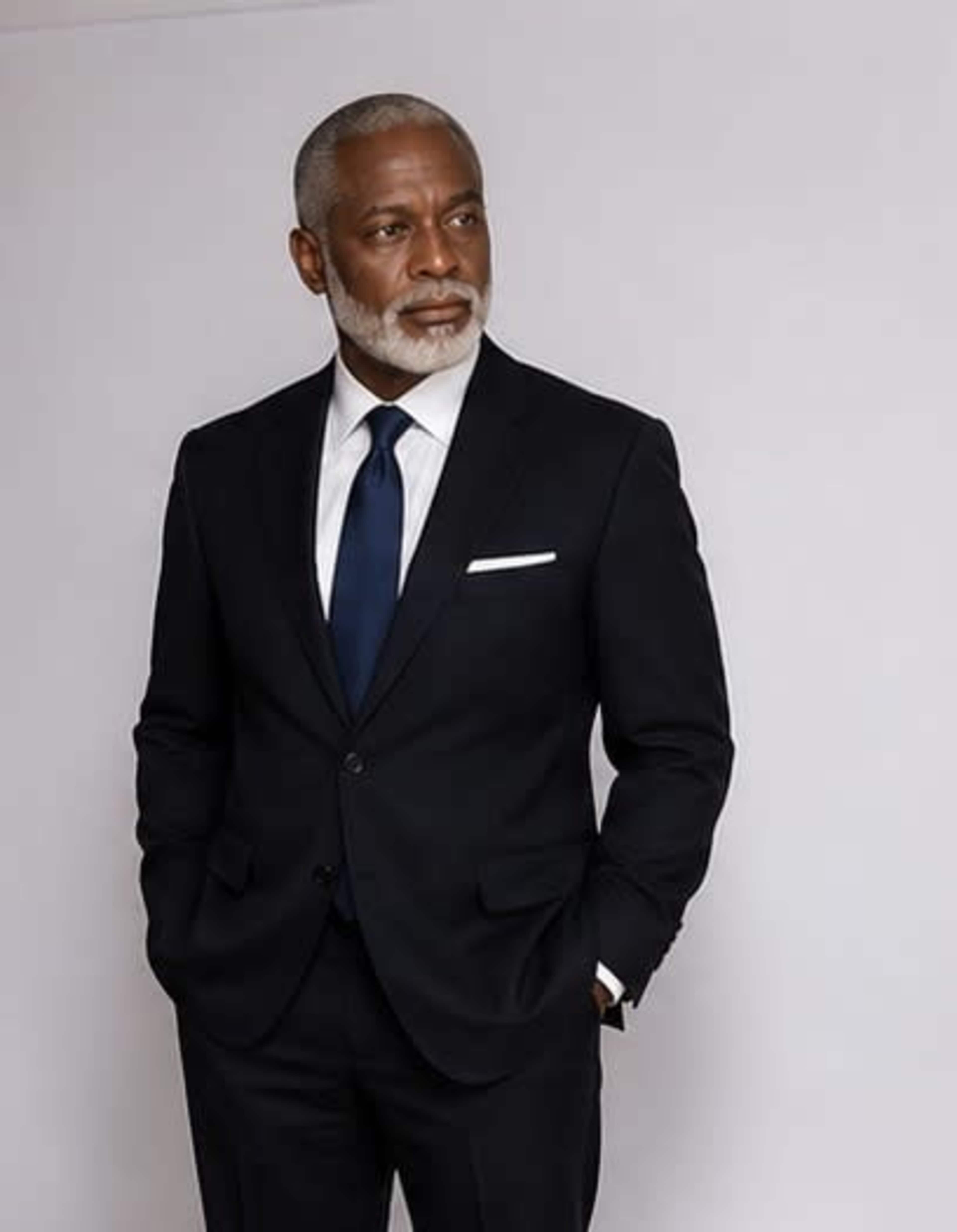 A professionally dressed man with a salt-and-pepper beard stands confidently against a plain background.