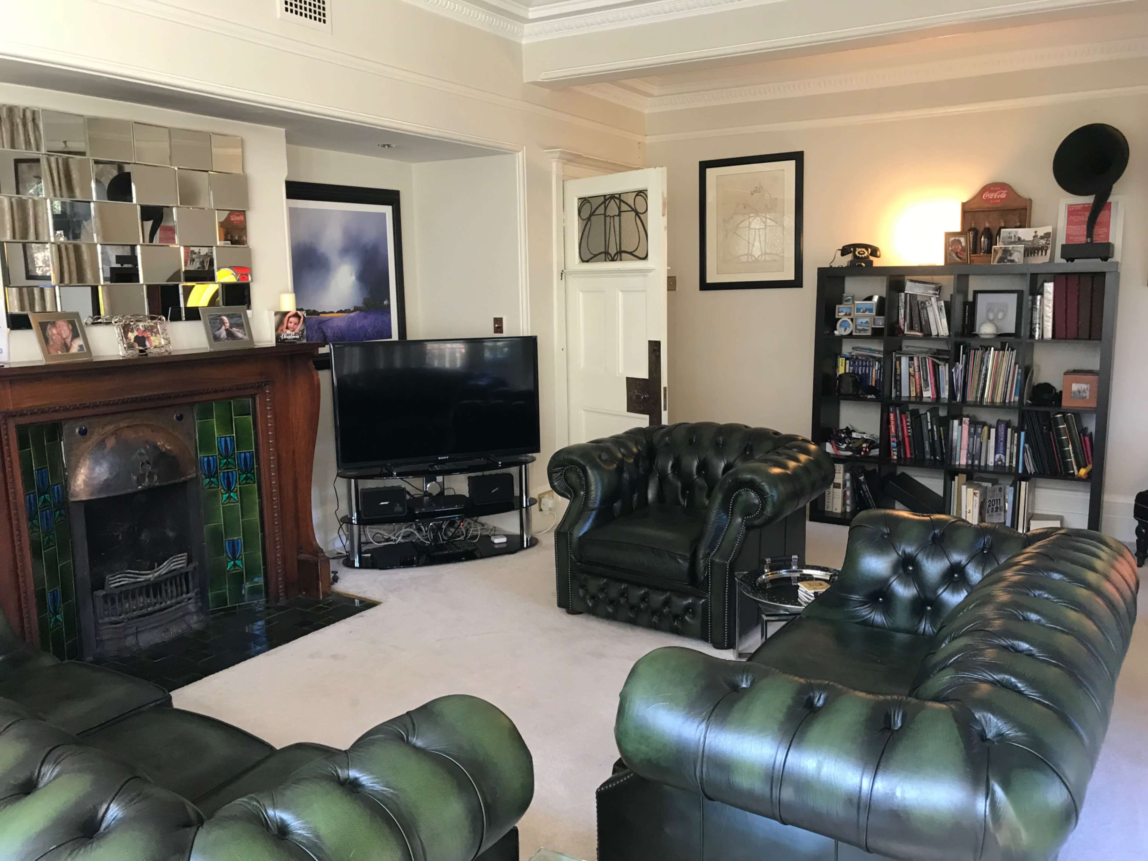 The living room features two green leather chesterfield sofas facing a television, with a decorative fireplace and bookshelves lining the walls.
