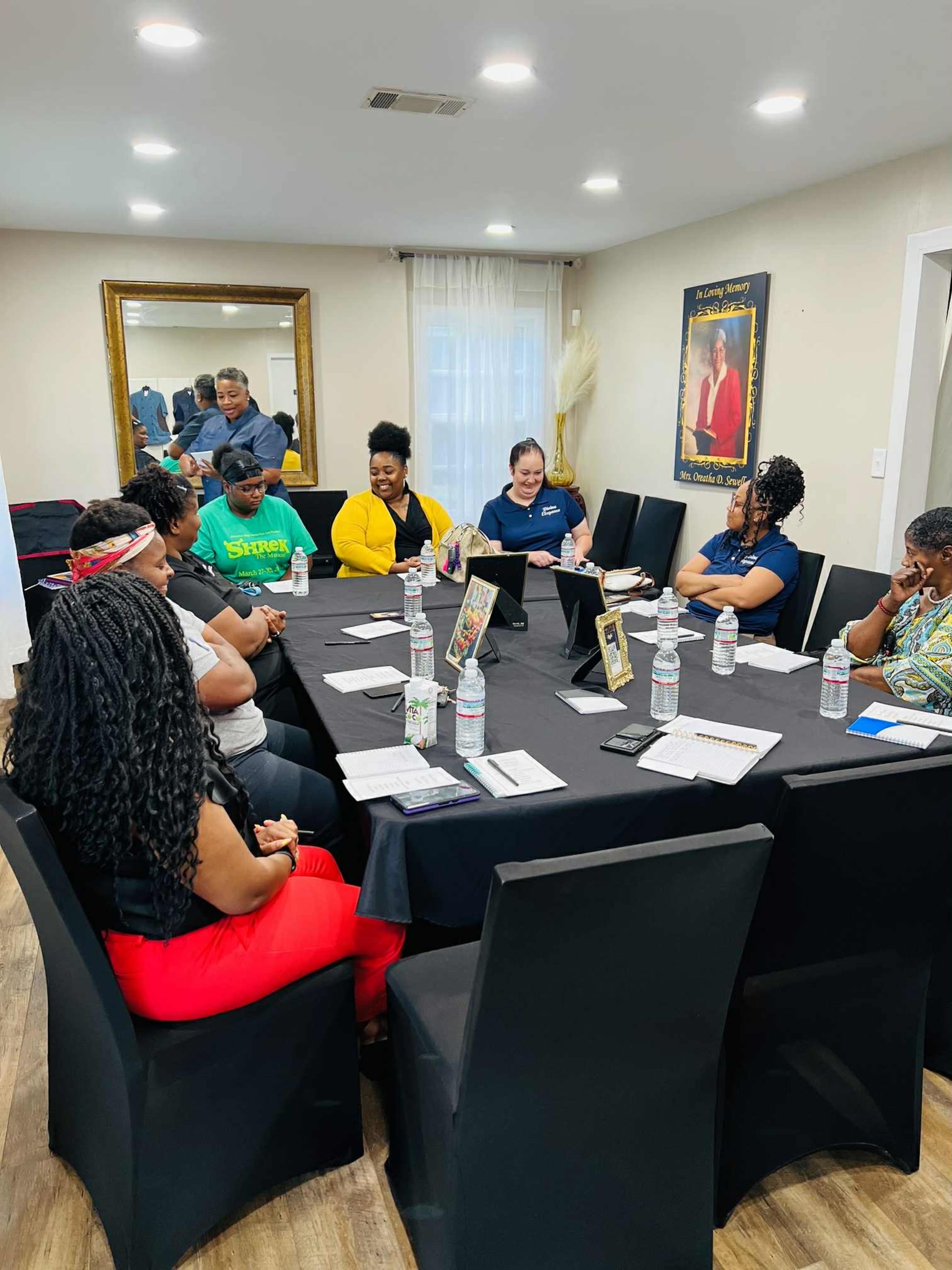 A group of people is seated around a rectangular table in a well-lit meeting room, engaging in a discussion with water bottles and notebooks in front of them.