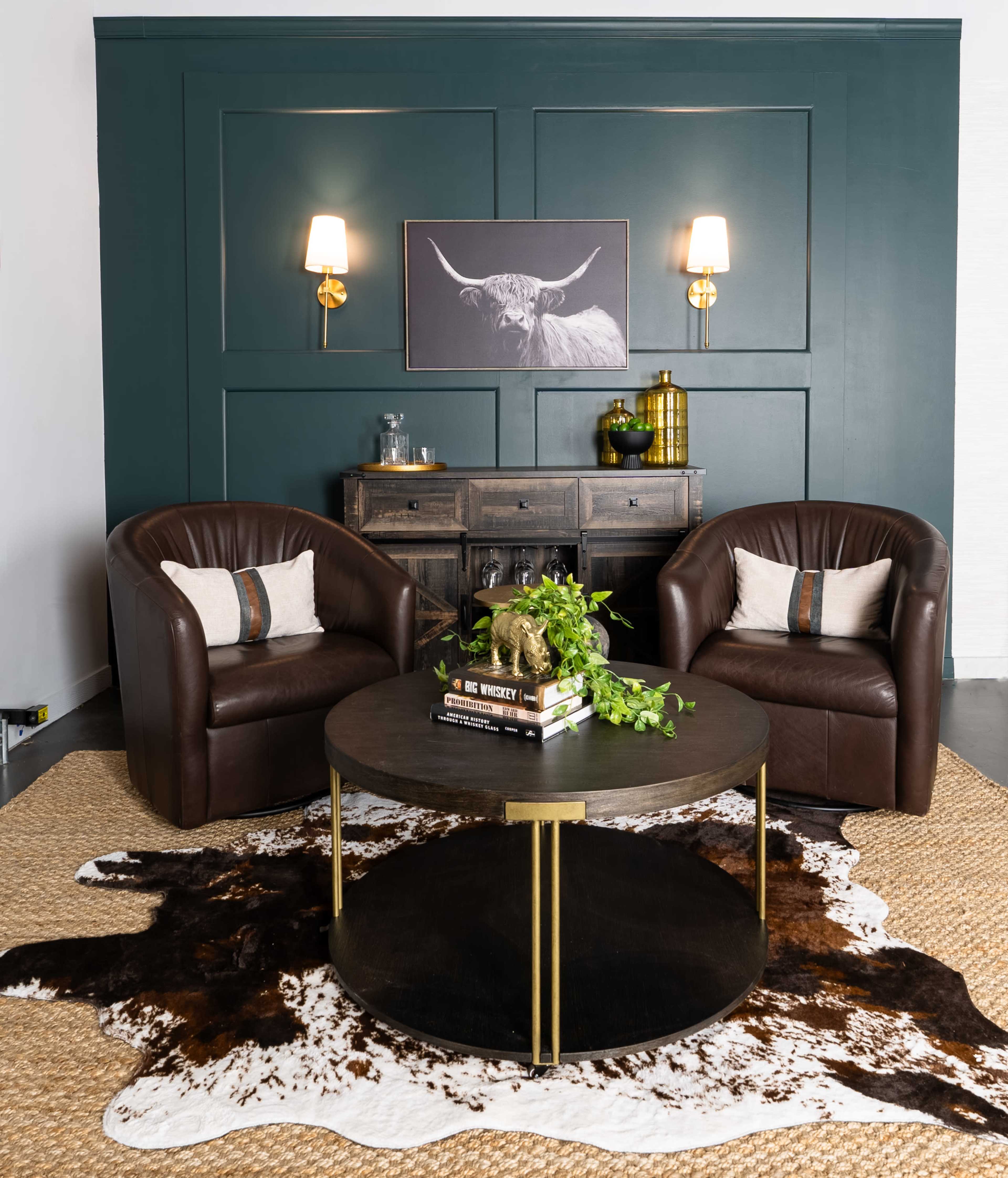 A cozy living area features two brown leather armchairs facing a round coffee table, surrounded by a cowhide rug, with a dark green wall and wall sconces in the background.
