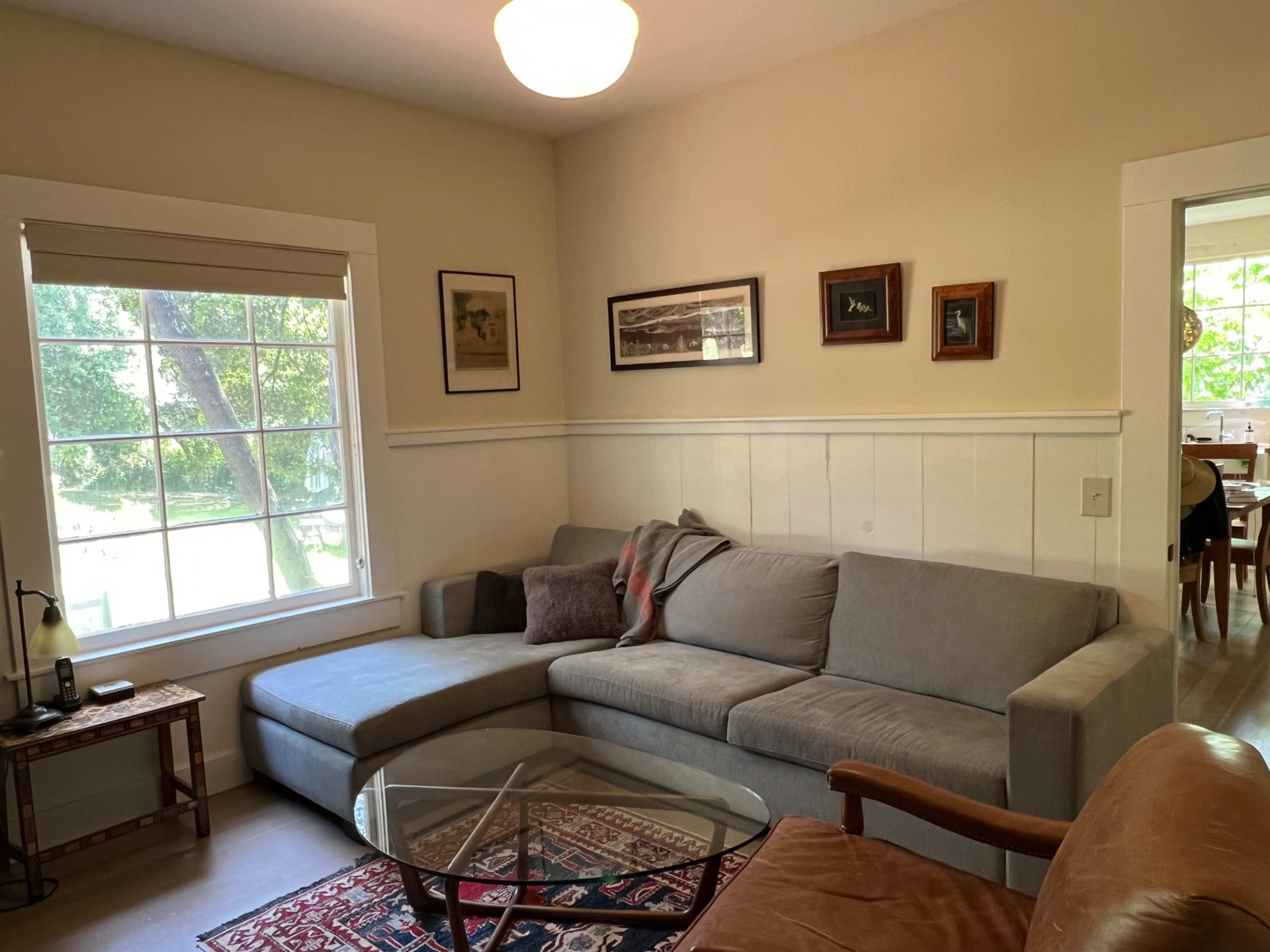 The image shows a cozy living room with a gray sectional sofa, a glass coffee table, and framed pictures on the walls, illuminated by natural light from a large window.
