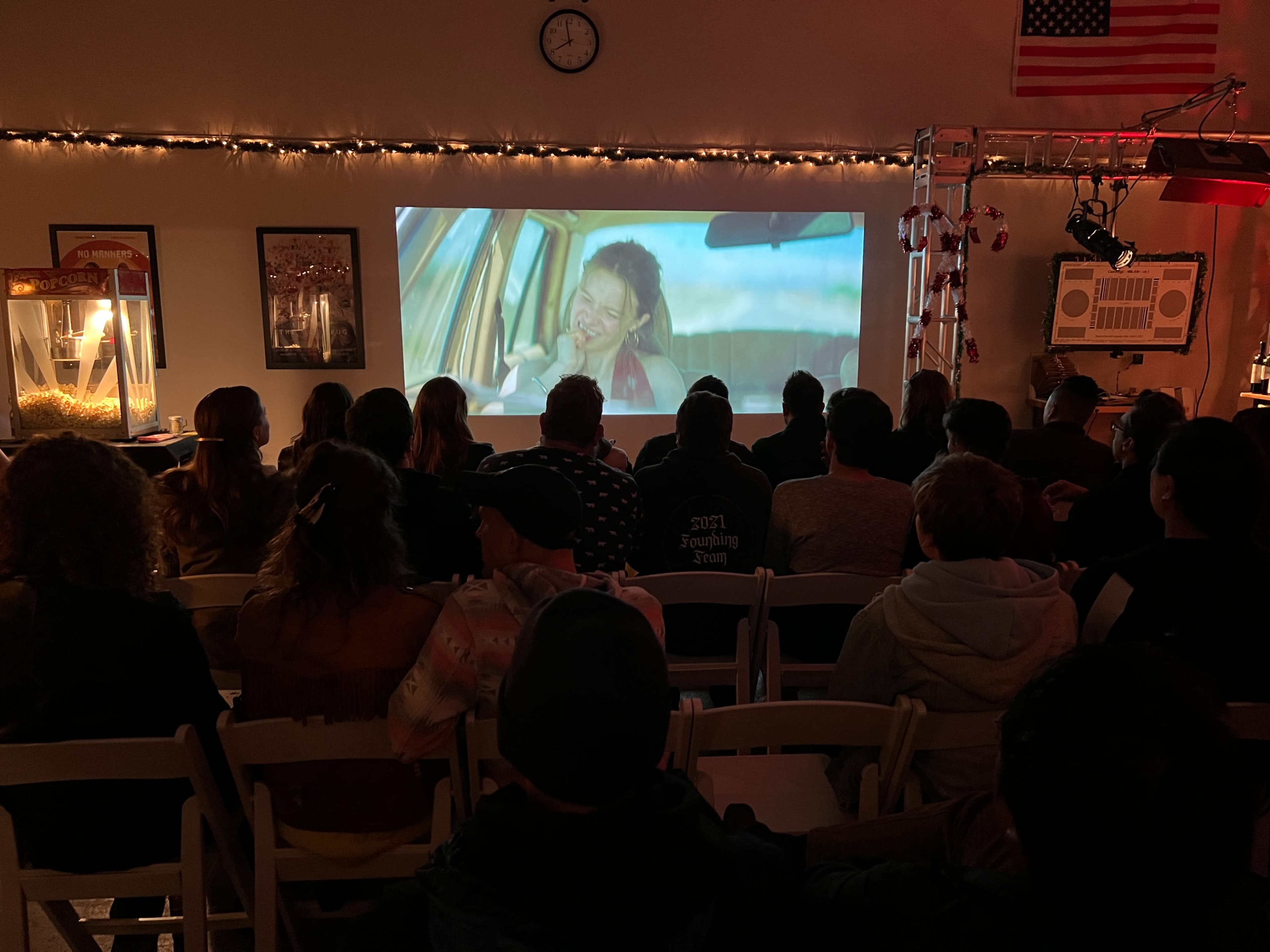 A group of people sit in chairs facing a screen displaying a scene from a movie in a decorated room.