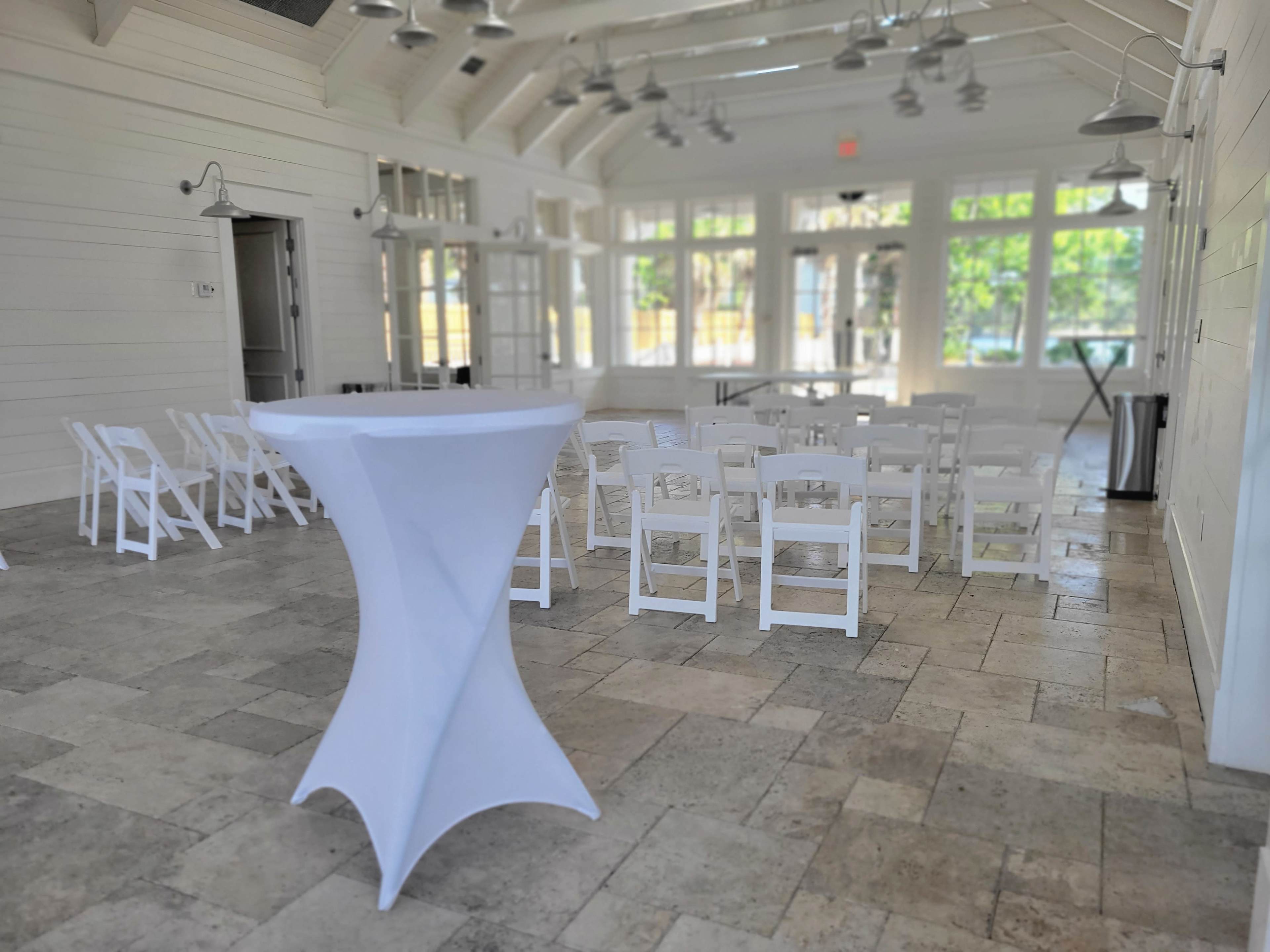 A brightly lit event space with white folding chairs arranged in rows facing a small stage, alongside a high-top table covered with a white tablecloth.