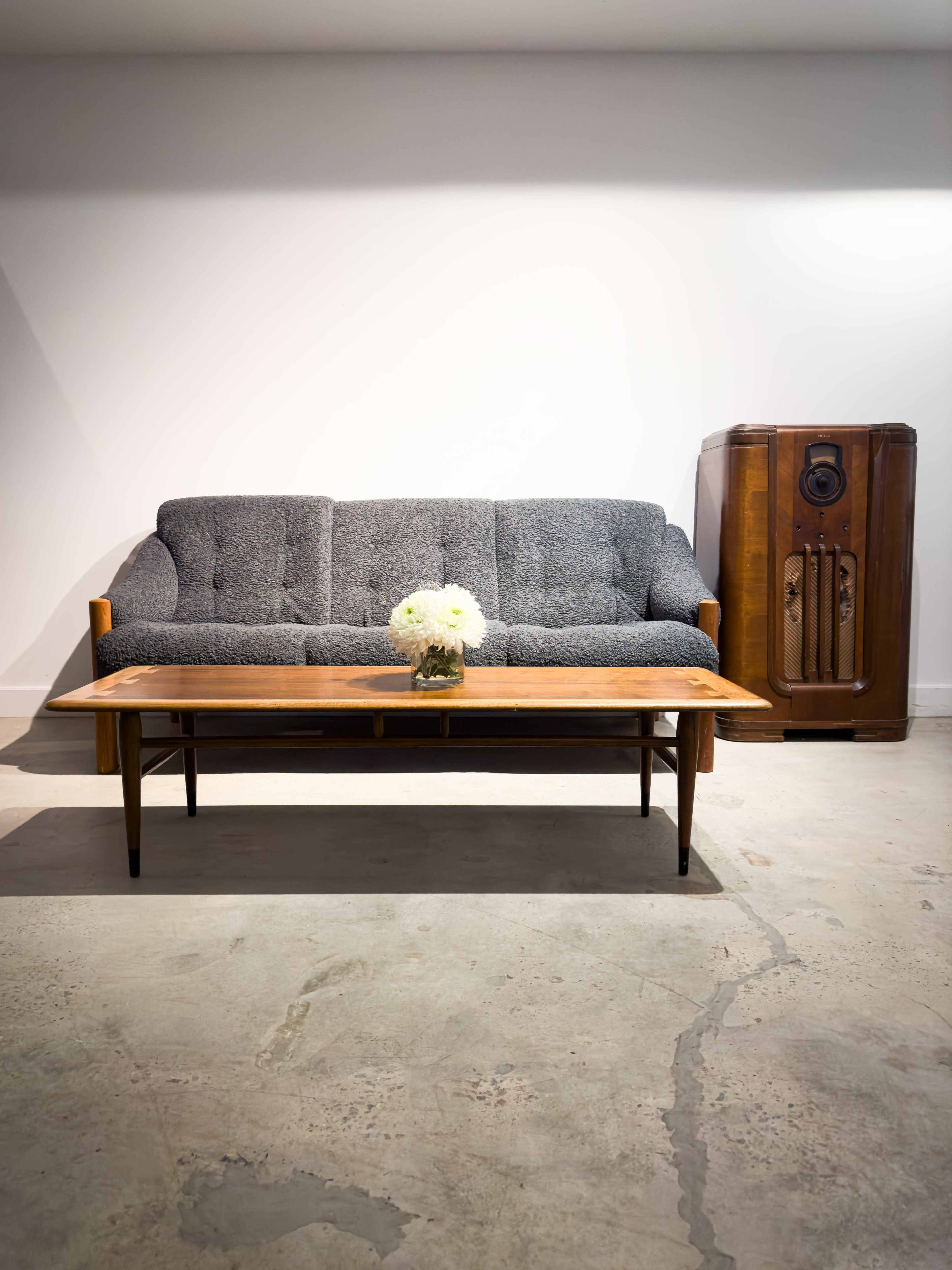 A gray upholstered sofa is positioned in front of a wooden coffee table, with a vase of white flowers on top, beside a vintage radio against a plain wall.