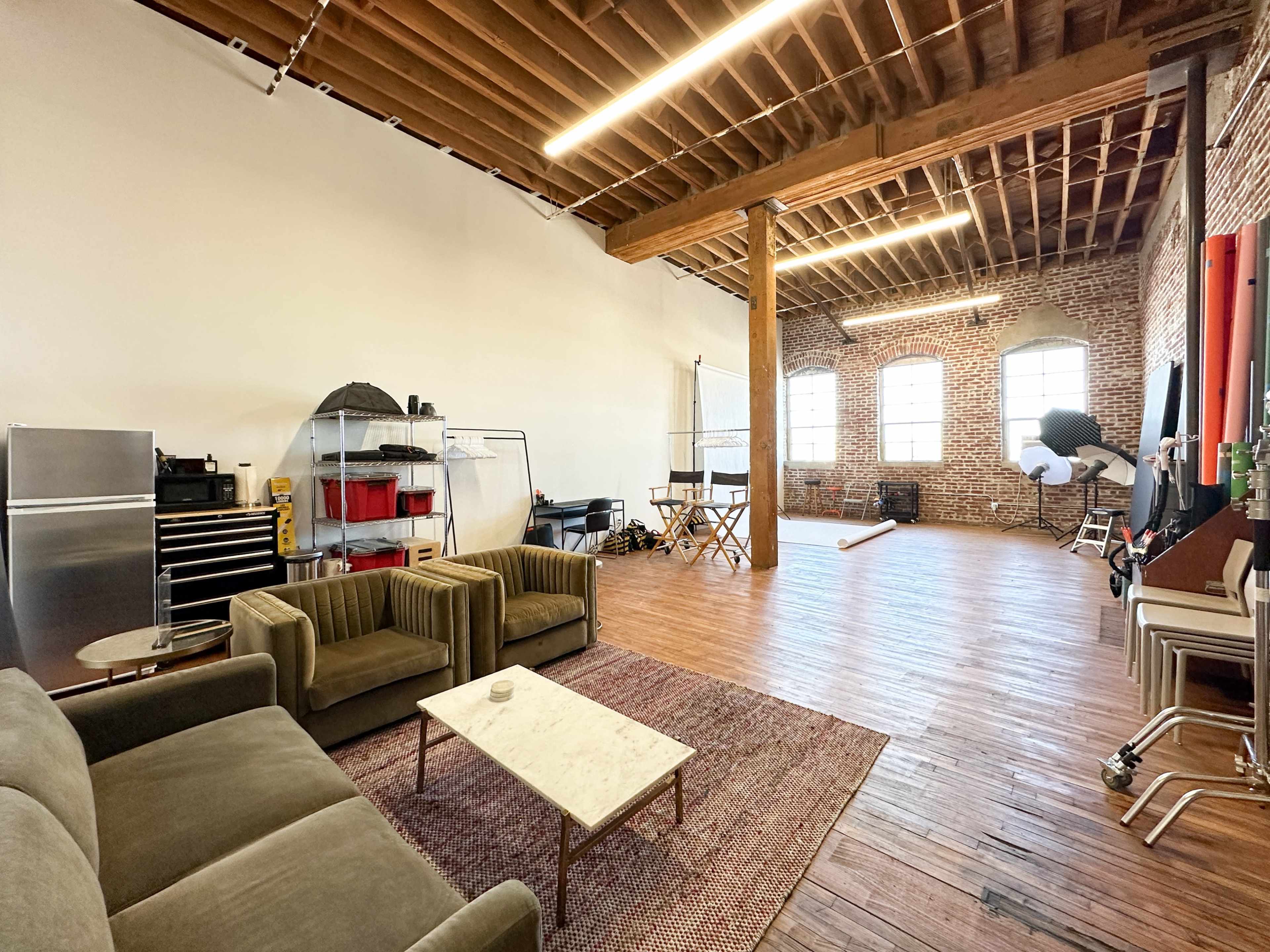 The image shows a spacious studio with exposed wooden beams, a brick wall, seating area with a coffee table, and various equipment set up for a photo shoot.