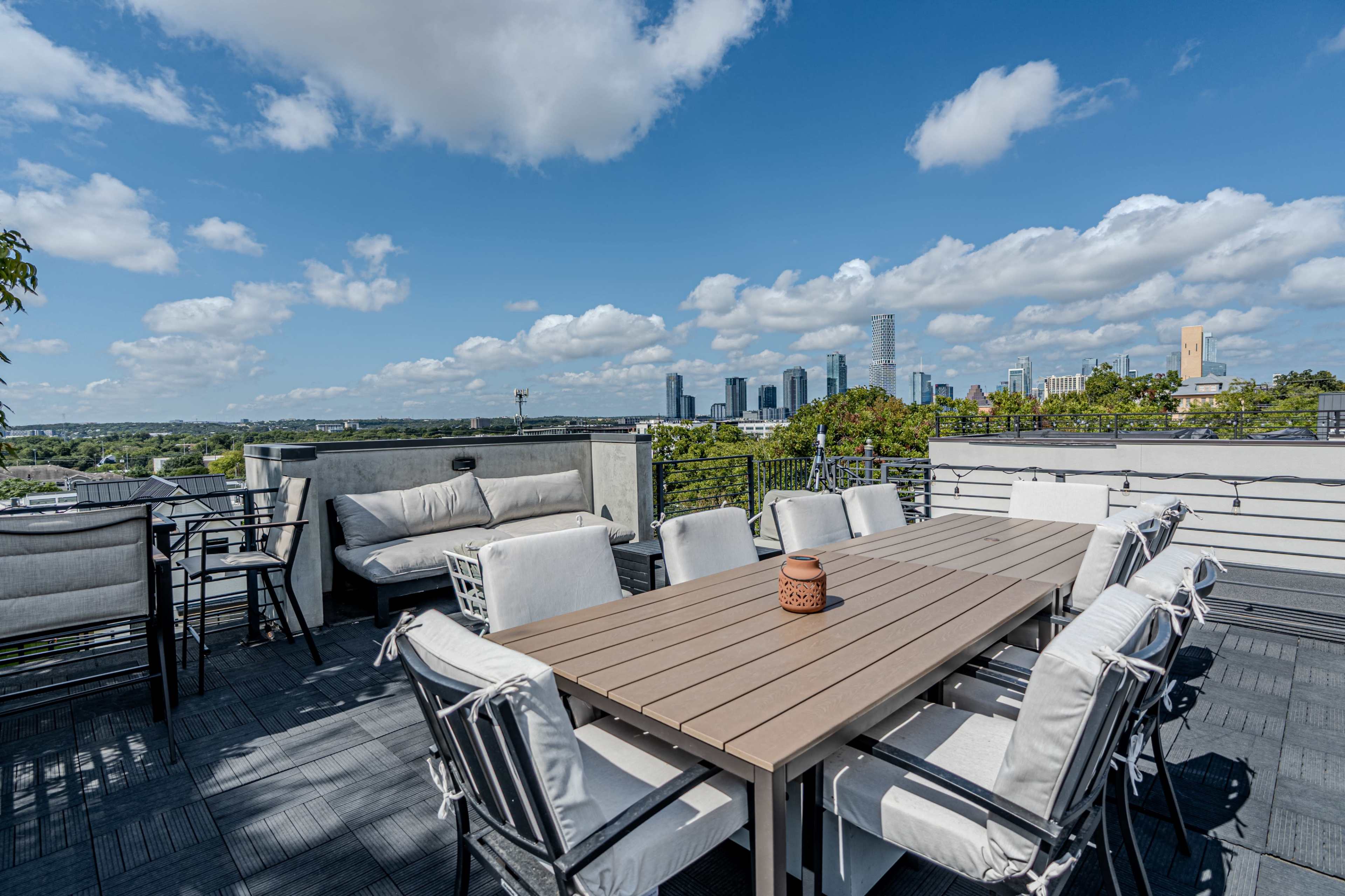 A rooftop patio with a large table and chairs overlooks a skyline filled with modern buildings under a partly cloudy sky.