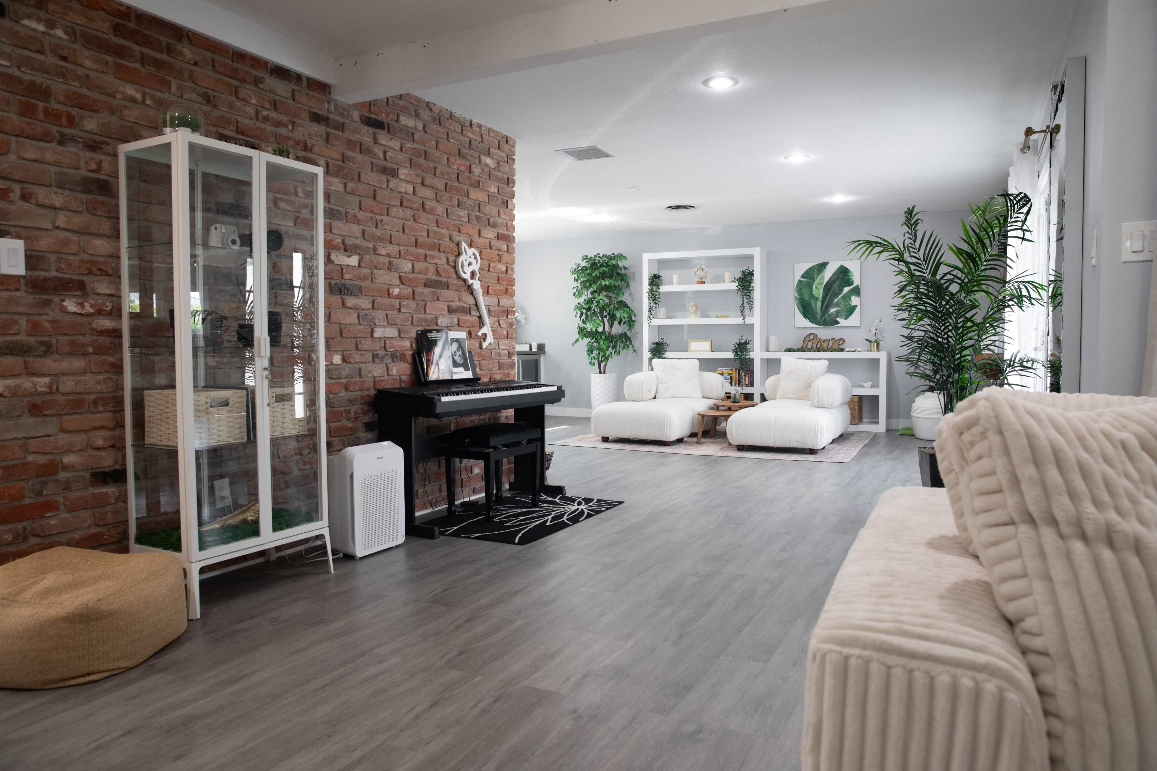 The image features a modern living space with a brick accent wall, a piano, white furniture, and greenery decor.