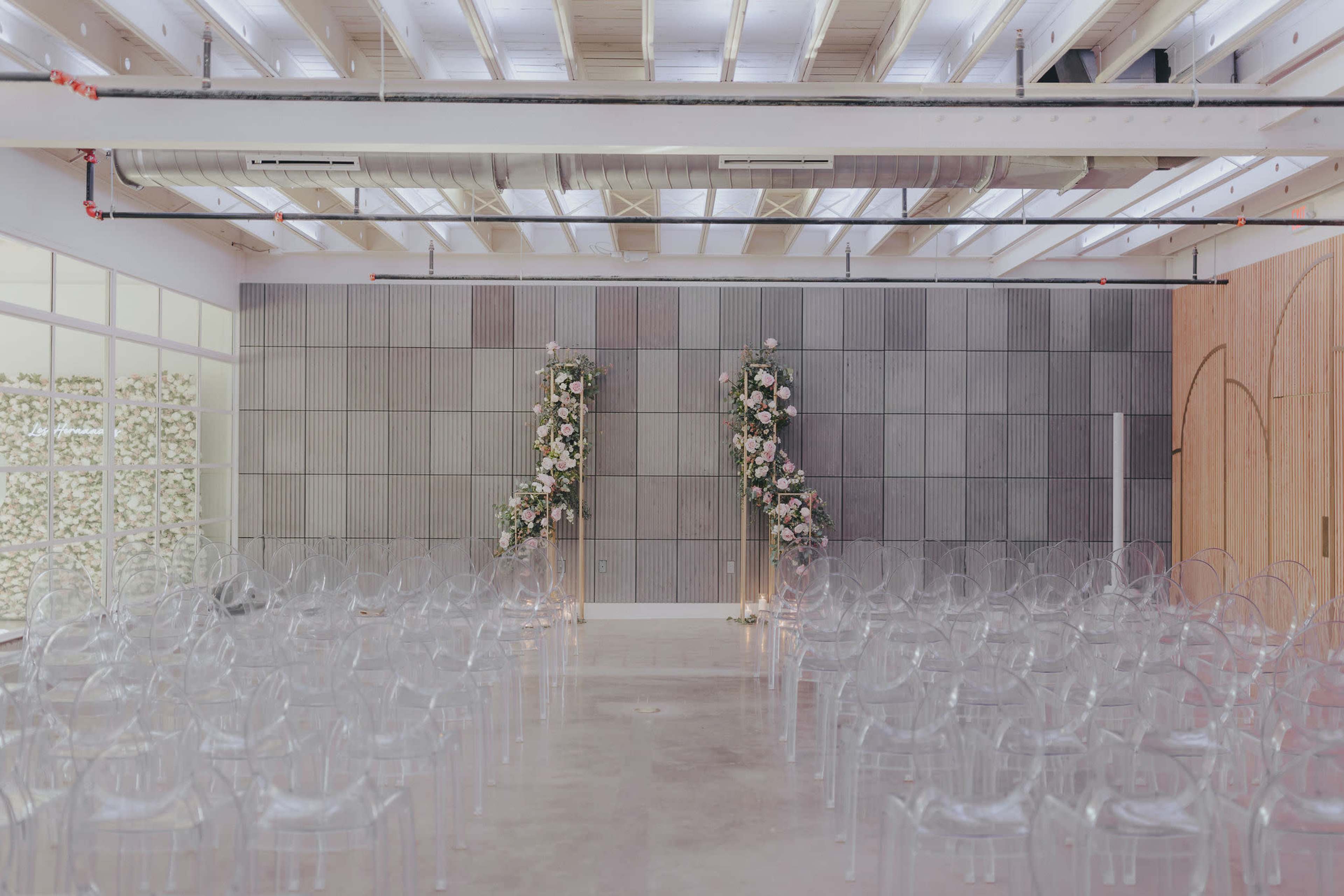 A modern wedding venue with transparent chairs arranged in rows facing a floral-adorned altar against a gray wall.