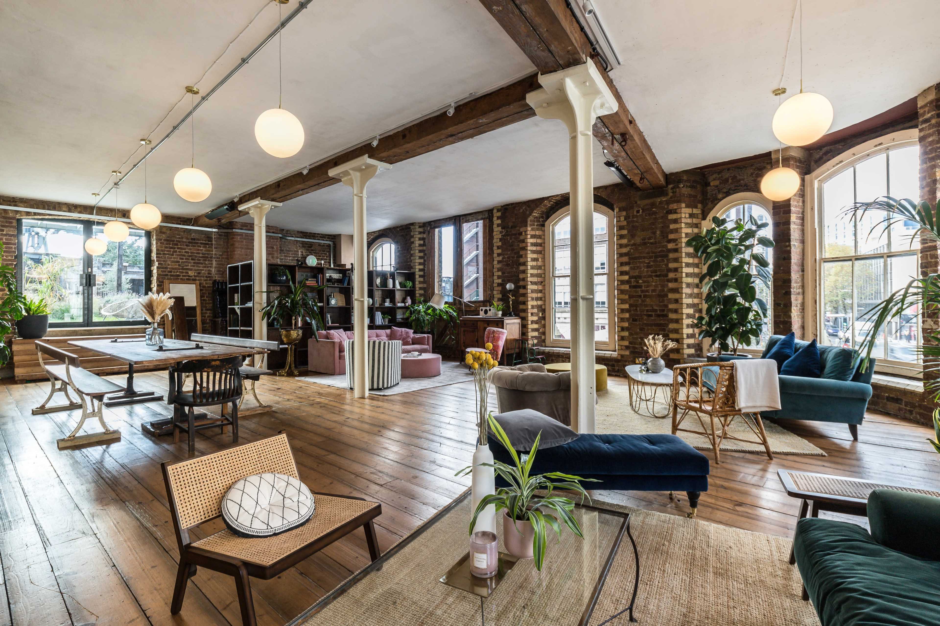 A spacious interior with wooden floors, large windows, and various seating and dining areas featuring plants and modern furniture.