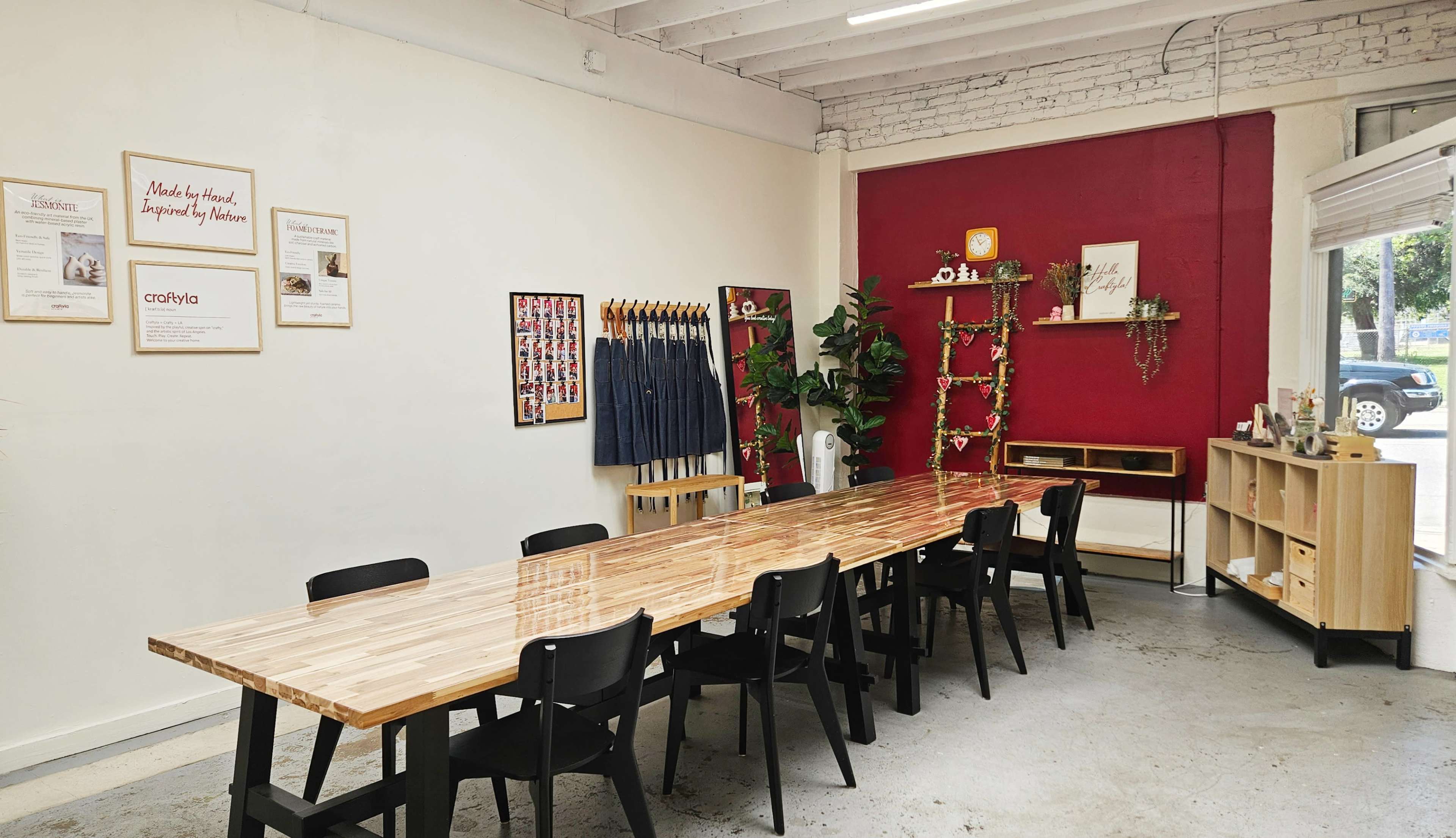 The image shows a spacious room featuring a long wooden table surrounded by black chairs, with a red accent wall and various decorative items displayed on nearby shelves.