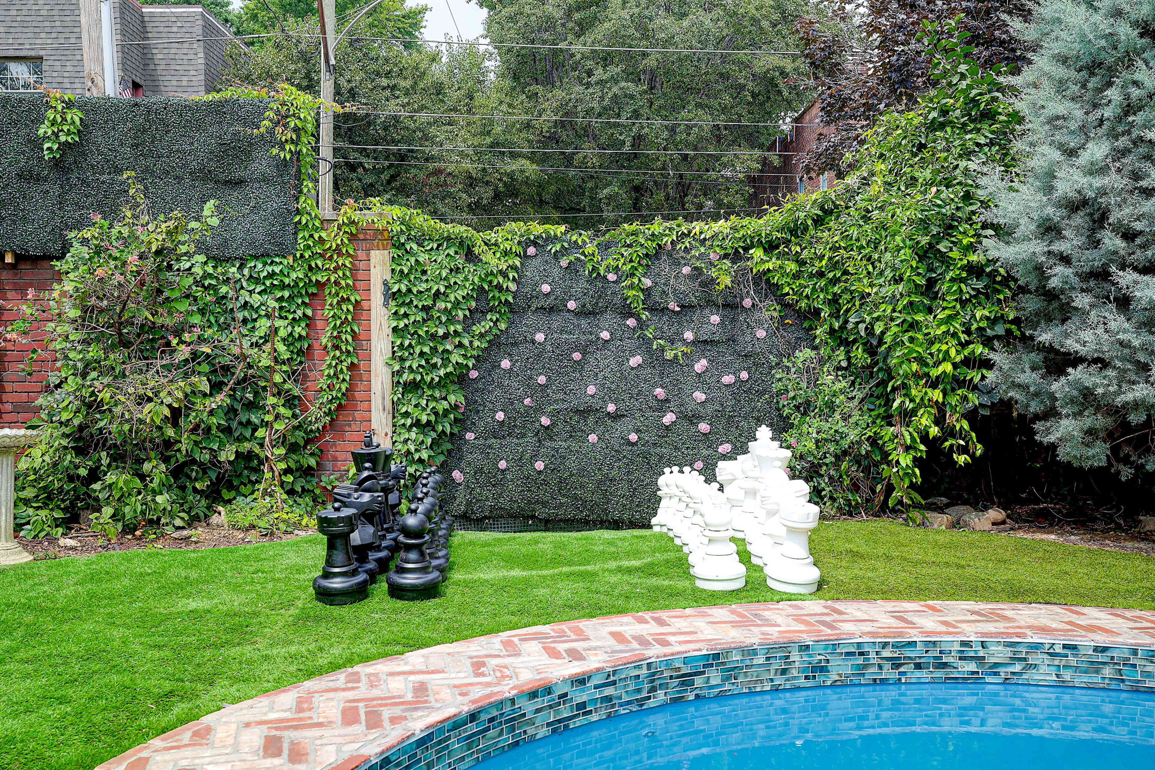 A large outdoor chess set with black and white pieces is arranged on the grass beside a pool, with a wall covered in greenery in the background.