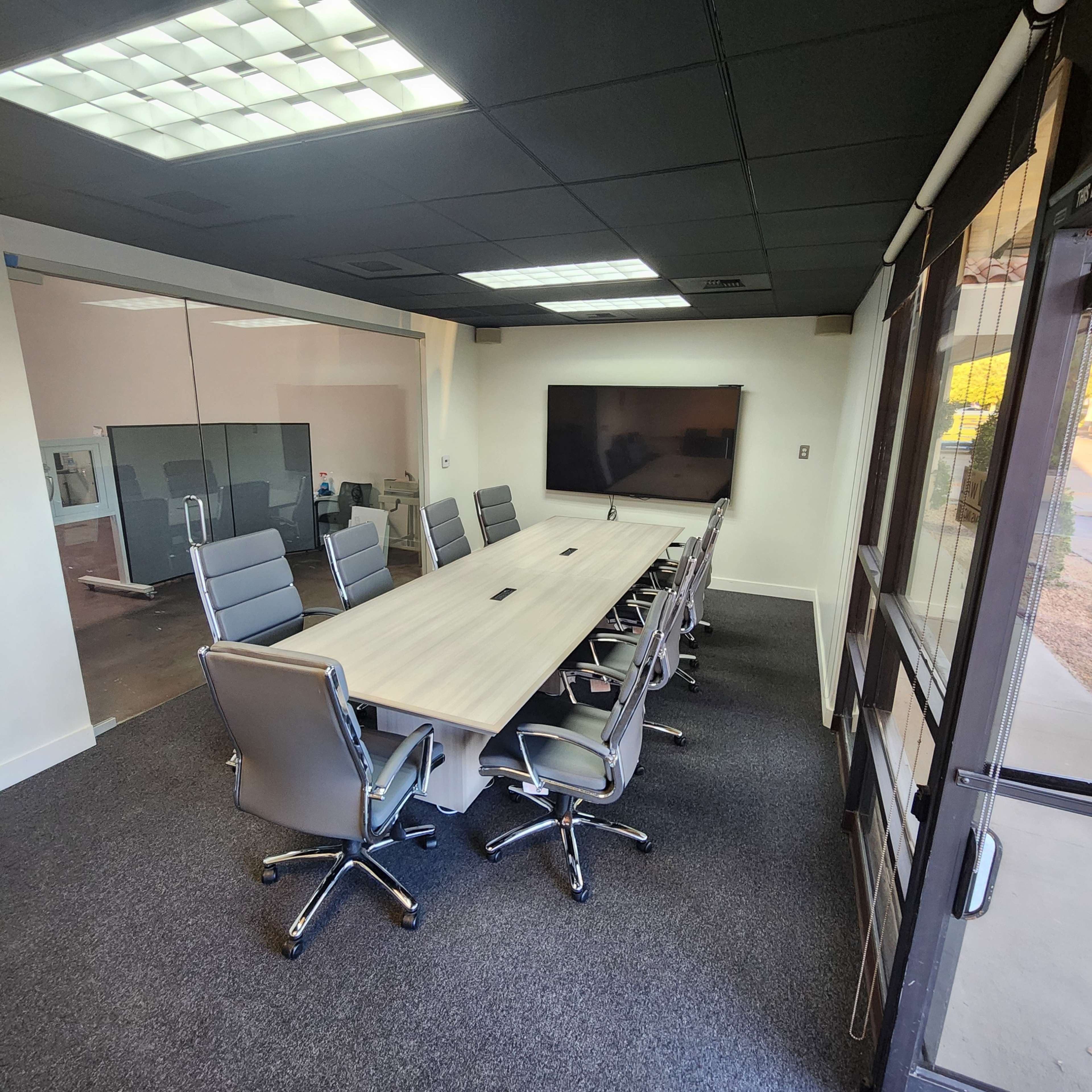 The image shows a modern conference room featuring a long table with chairs, a large screen on the wall, and glass walls providing visibility to an adjacent space.