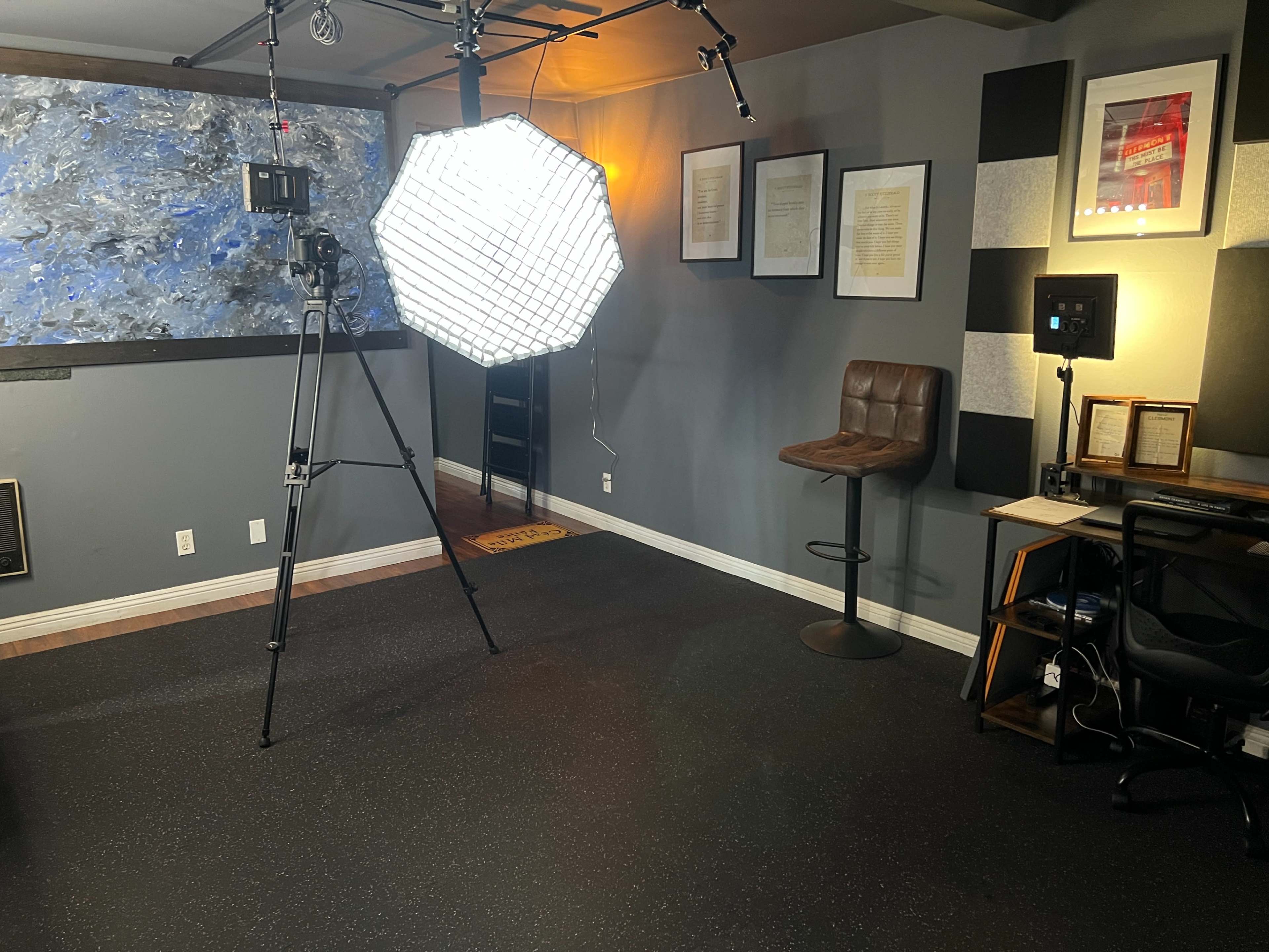 A studio space with a camera on a tripod, a large softbox light, a high stool, framed artwork on the wall, and a desk with a chair and equipment.