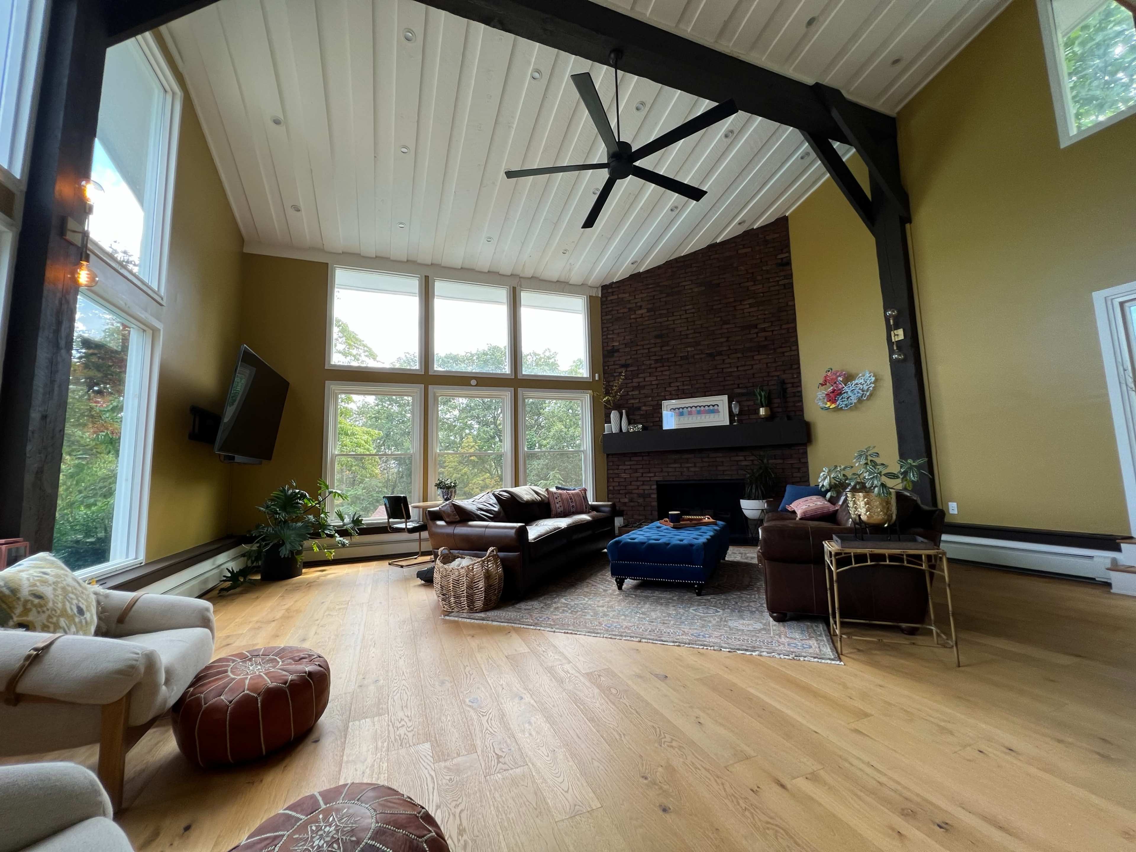 A spacious living room with large windows, a brick fireplace, and decorative furniture, including a blue ottoman and a patterned rug.