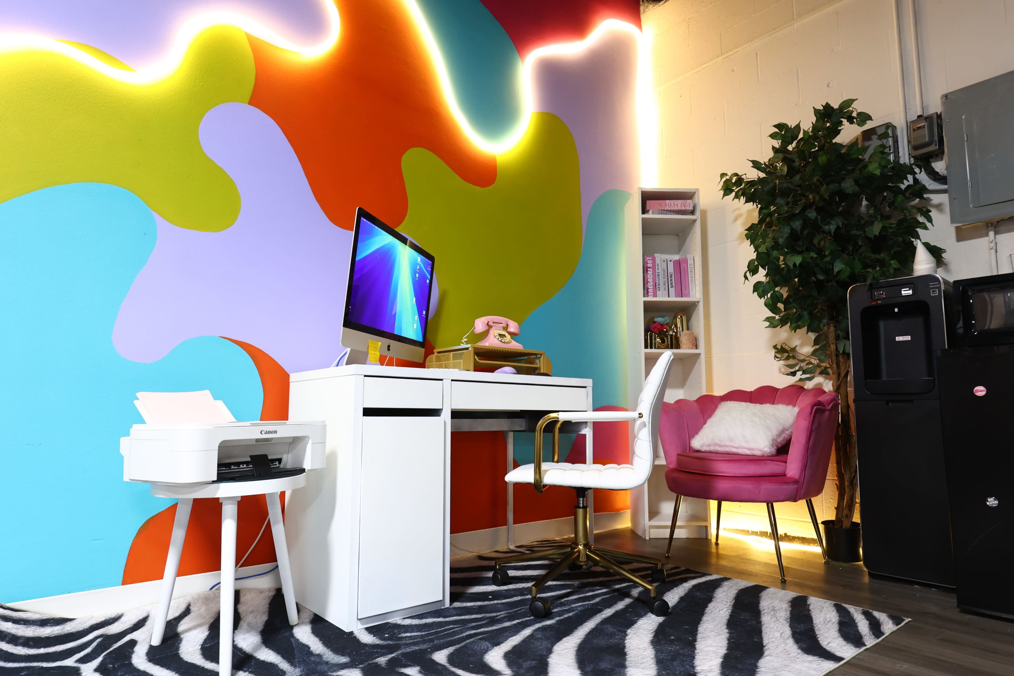 The image shows a colorful office space with a modern desk and computer, a pink chair, and a vibrant, abstract wall design.