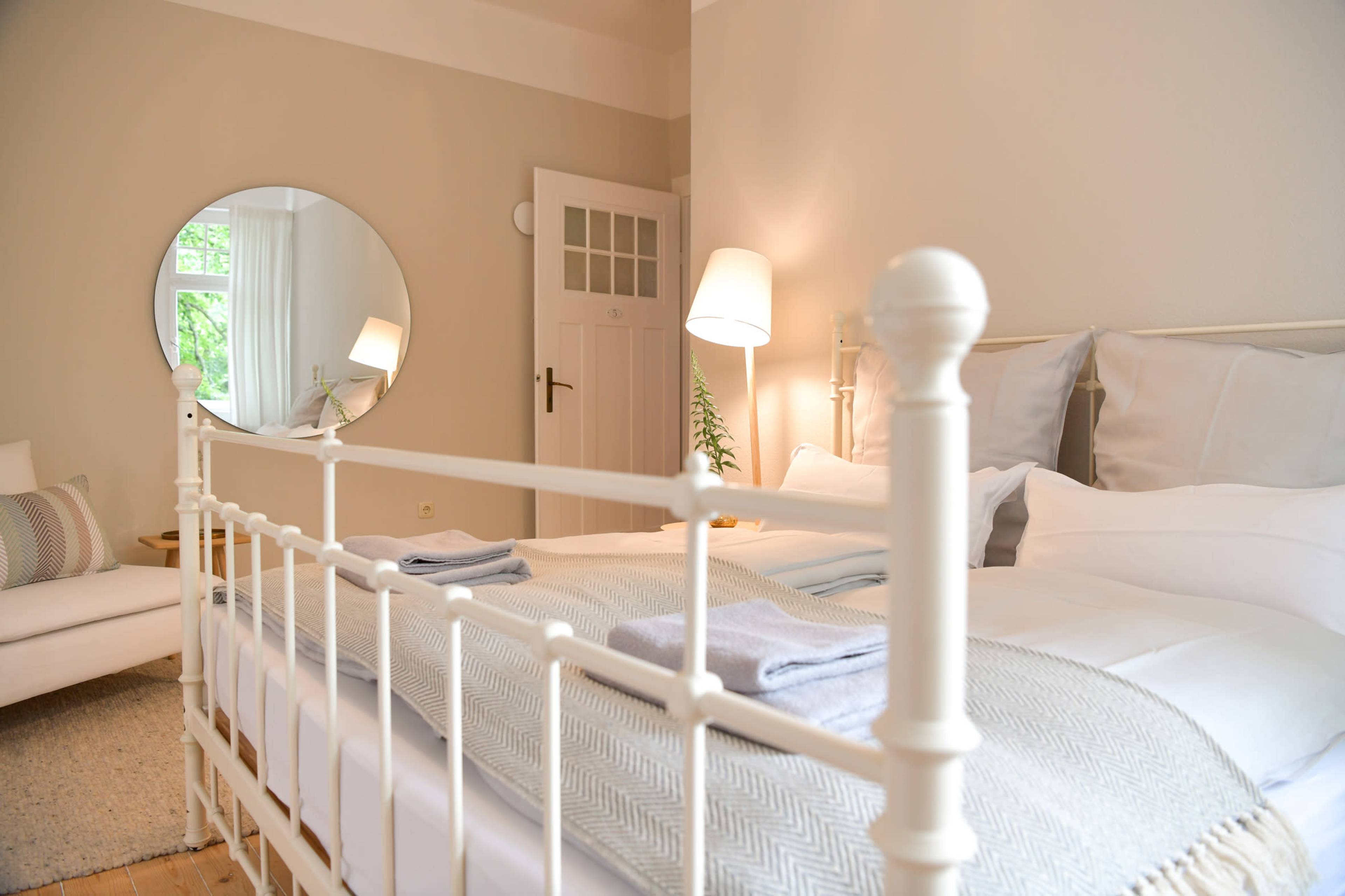 A neatly arranged bedroom featuring a white metal bed, soft linens, a round mirror, and a cozy seating area.