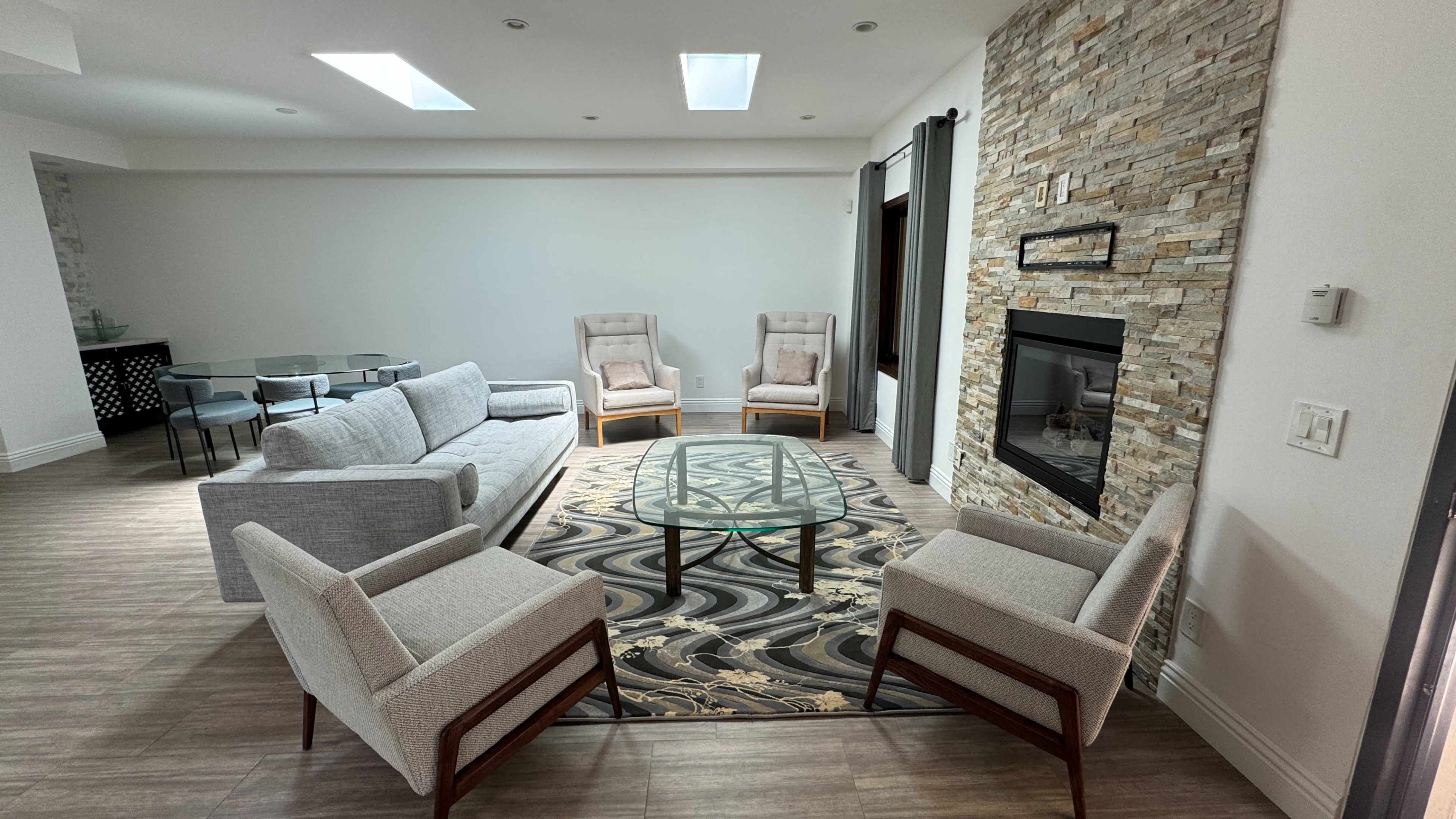 The image shows a modern living room featuring a gray sectional sofa, two armchairs, a glass coffee table, and a stone fireplace, with natural light coming from the skylights.