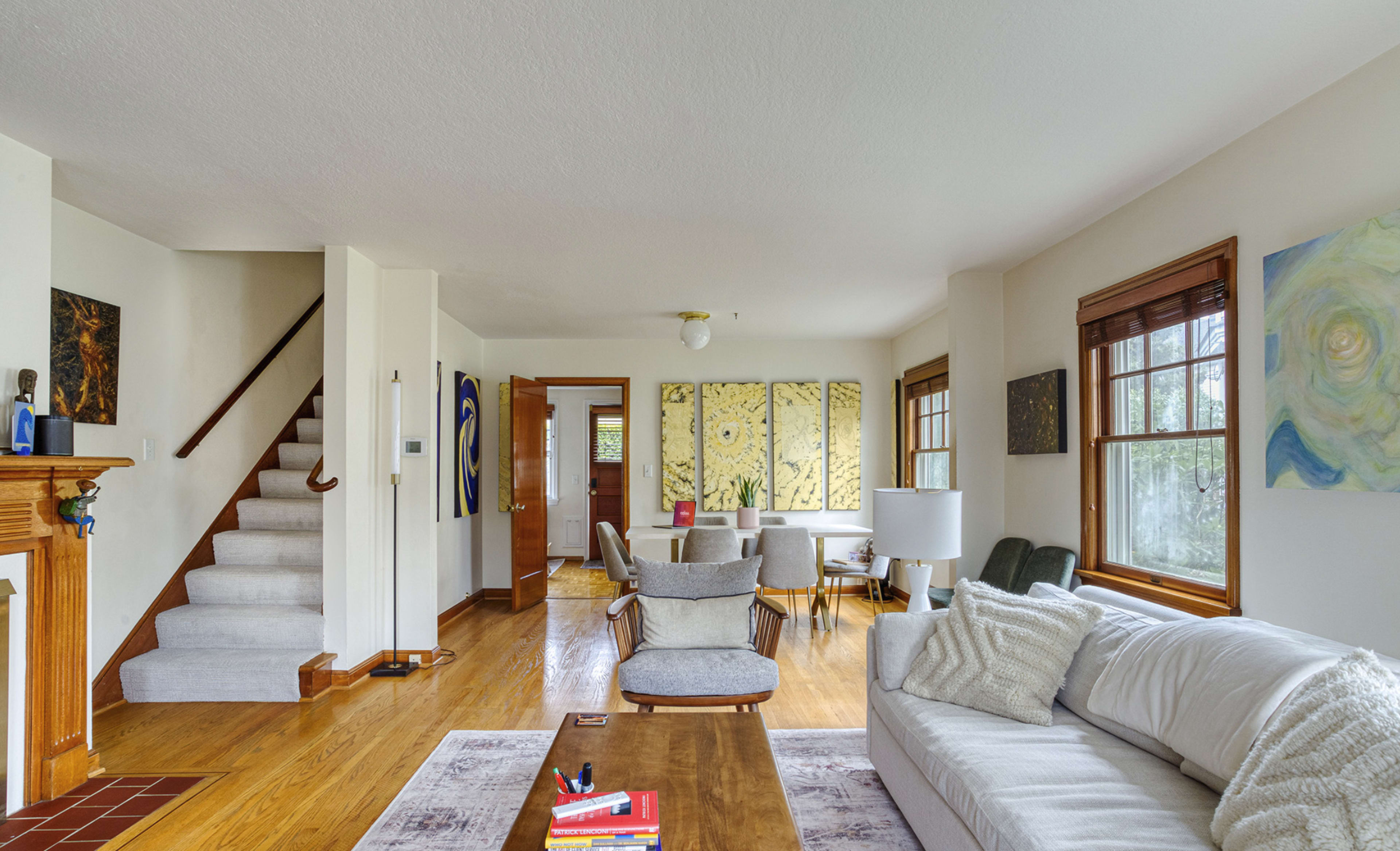 The image shows a bright living room with a white sofa, a wooden coffee table, and a staircase leading to an upper level.