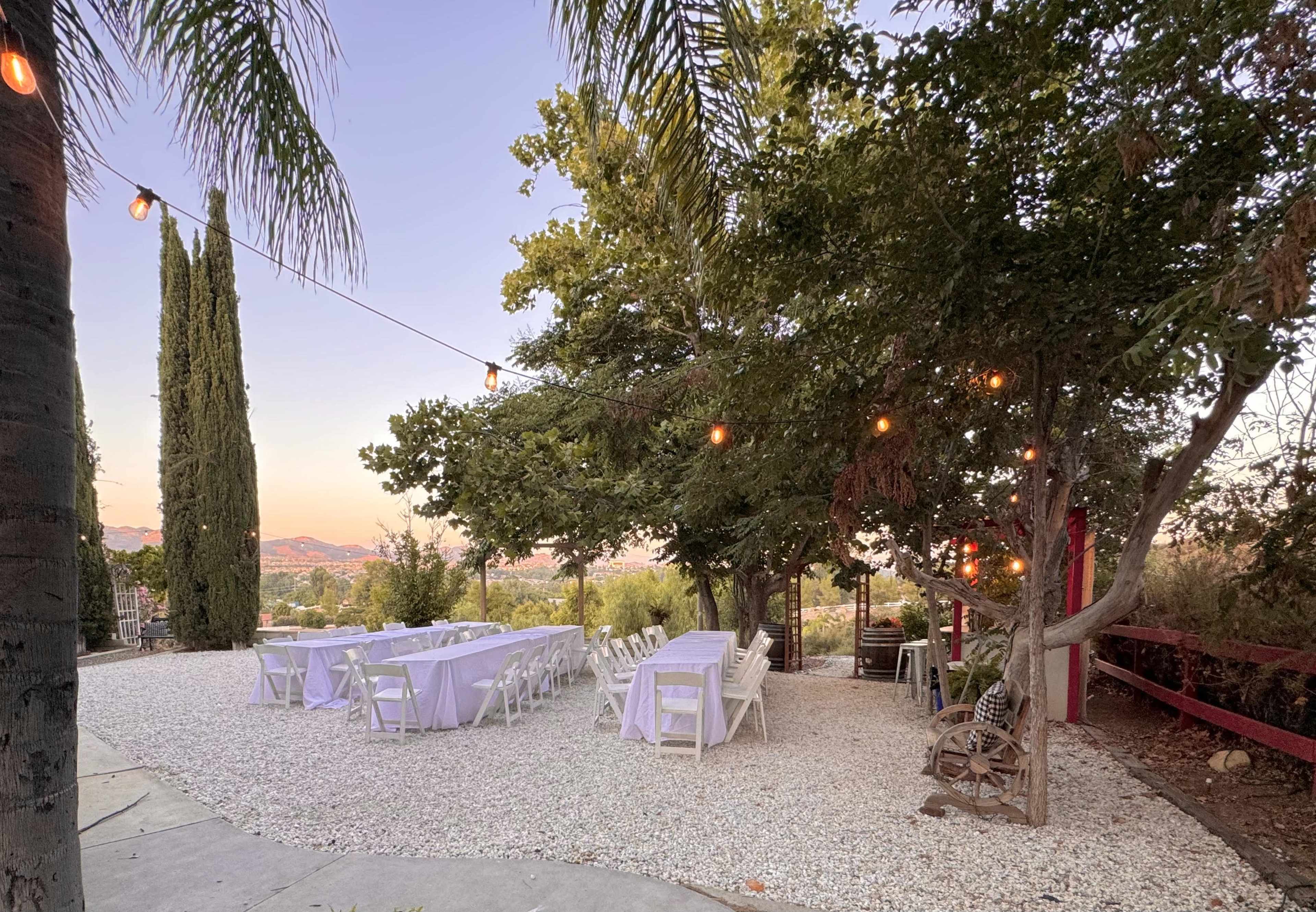 The scene depicts a landscaped outdoor area set for a gathering, featuring tables covered with white cloths, string lights hanging above, and a backdrop of greenery and distant hills.