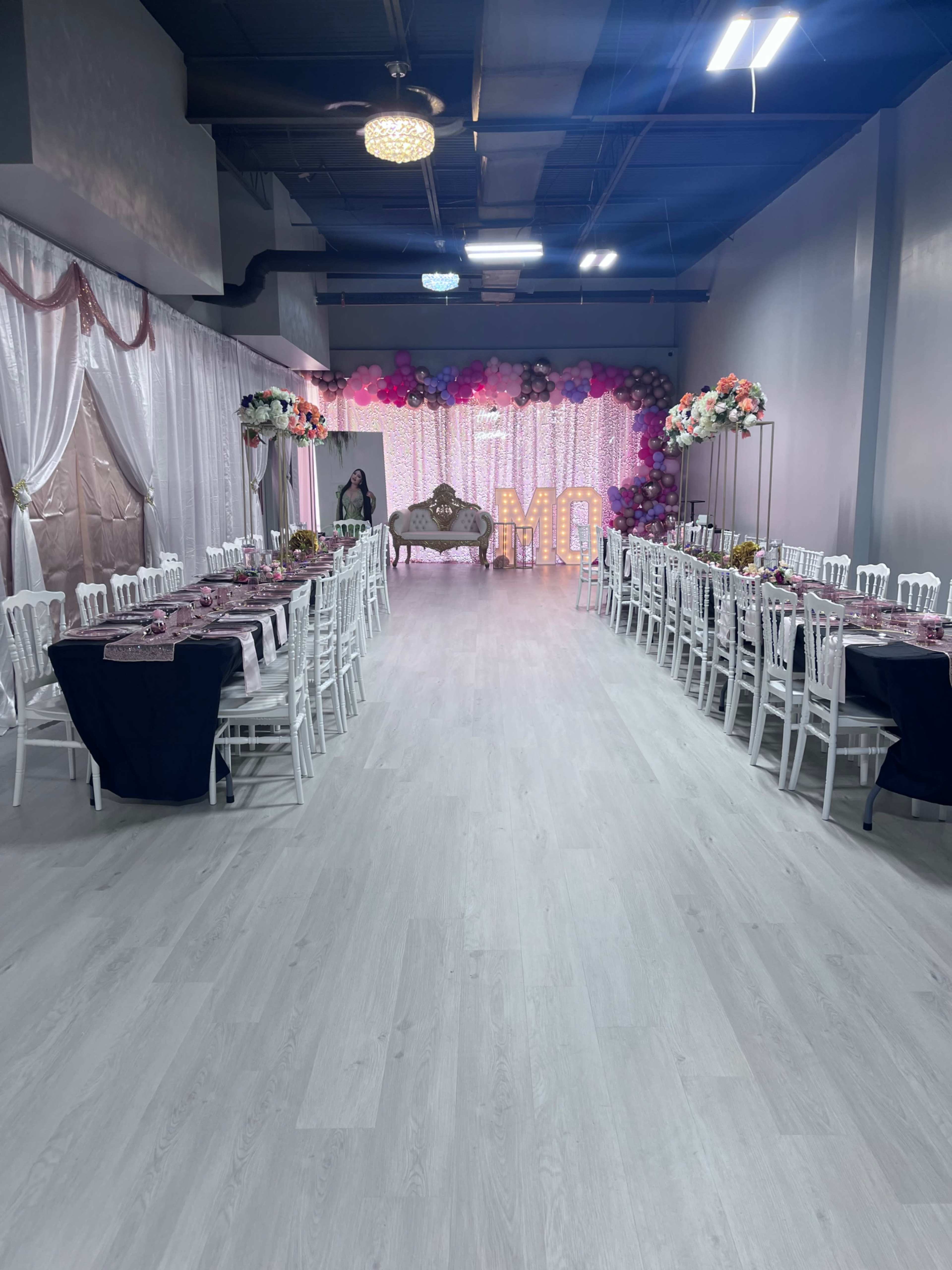 A decorated event space featuring long tables set for a gathering, with a backdrop of lights and colorful balloon arrangements at one end.