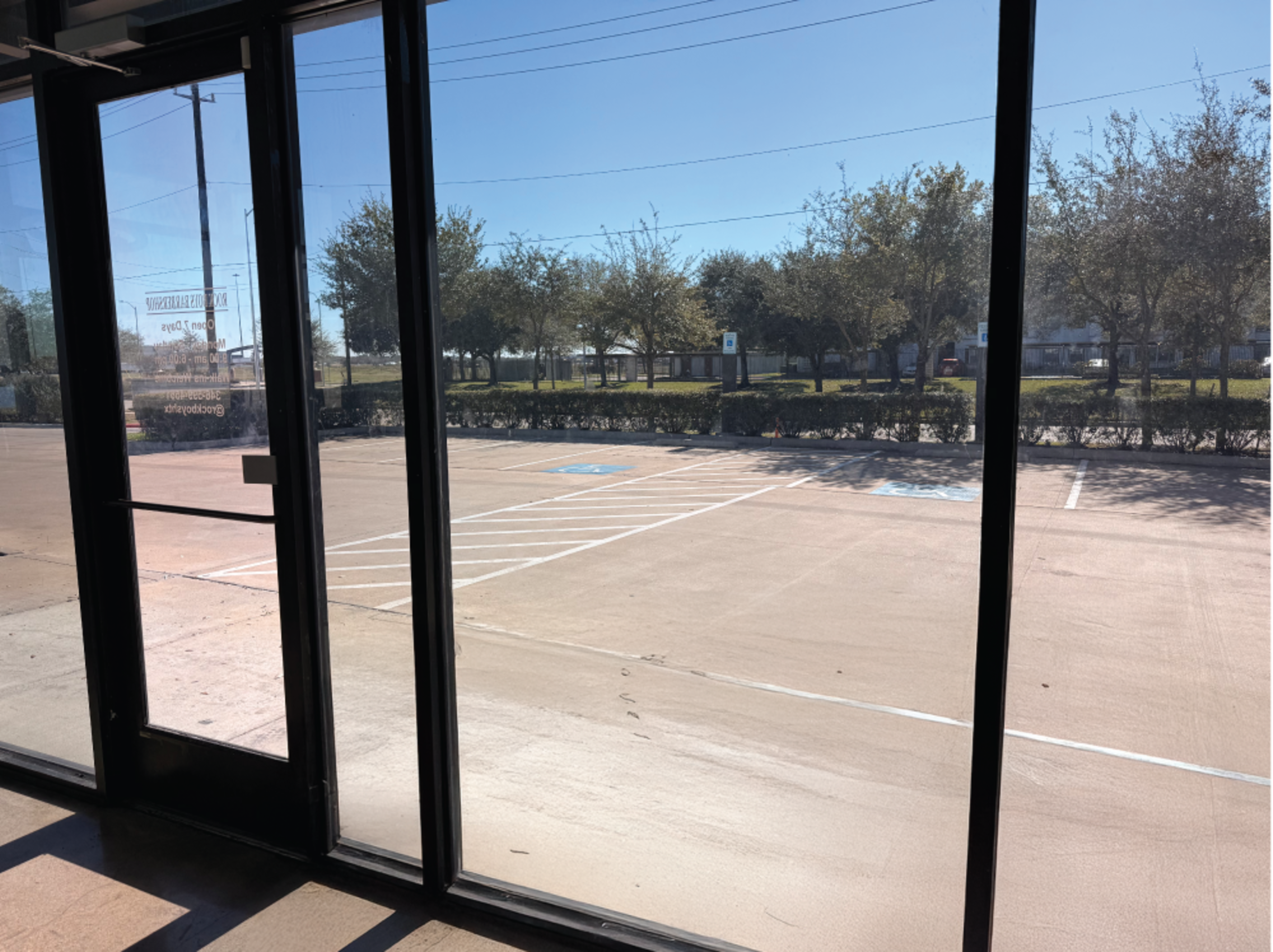 The image shows an empty parking lot viewed through the glass doors of a building, with trees lining the background and clear blue skies visible.
