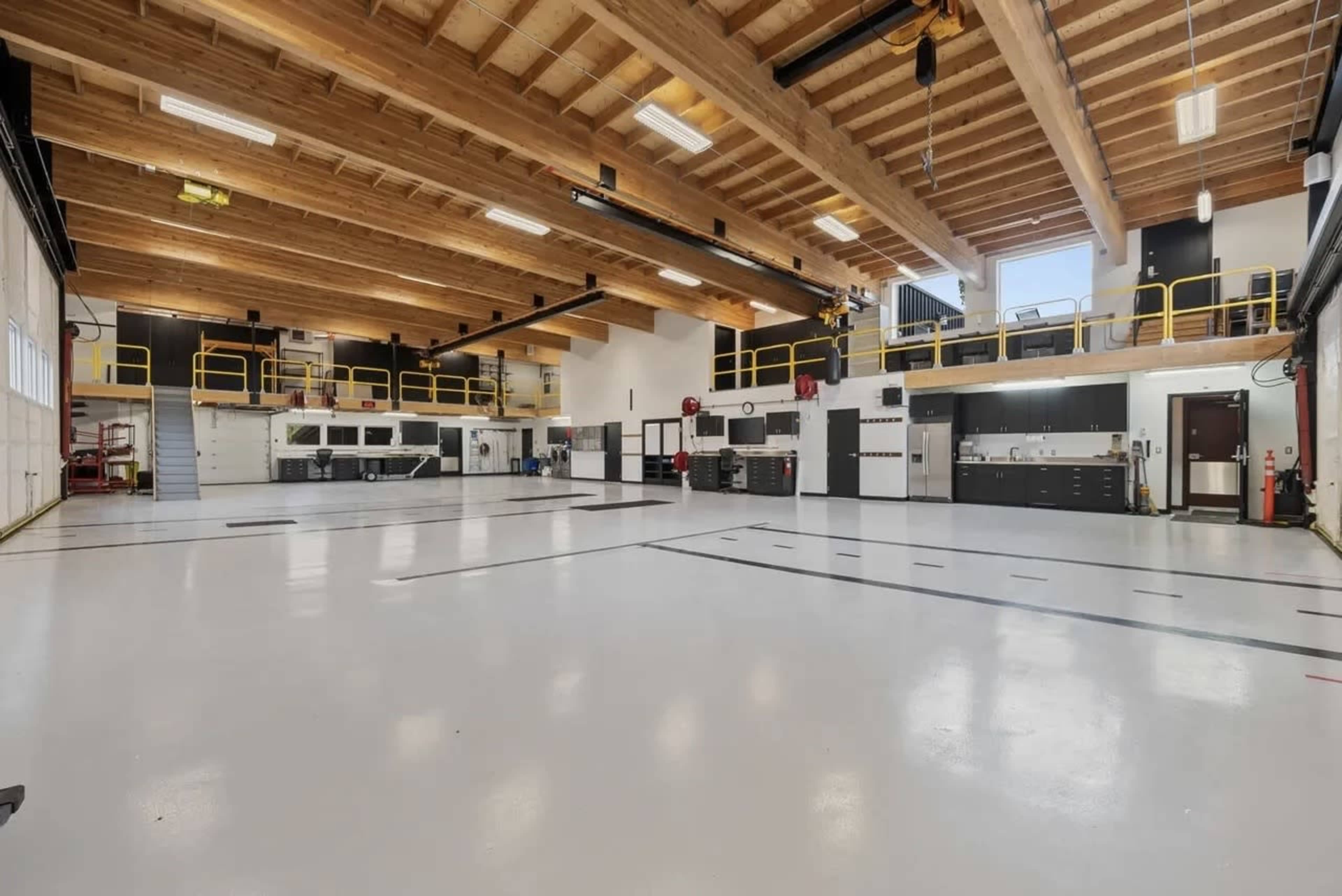 The image shows a spacious, modern workshop with high wooden ceilings, numerous garage doors, and organized workstations along the walls.