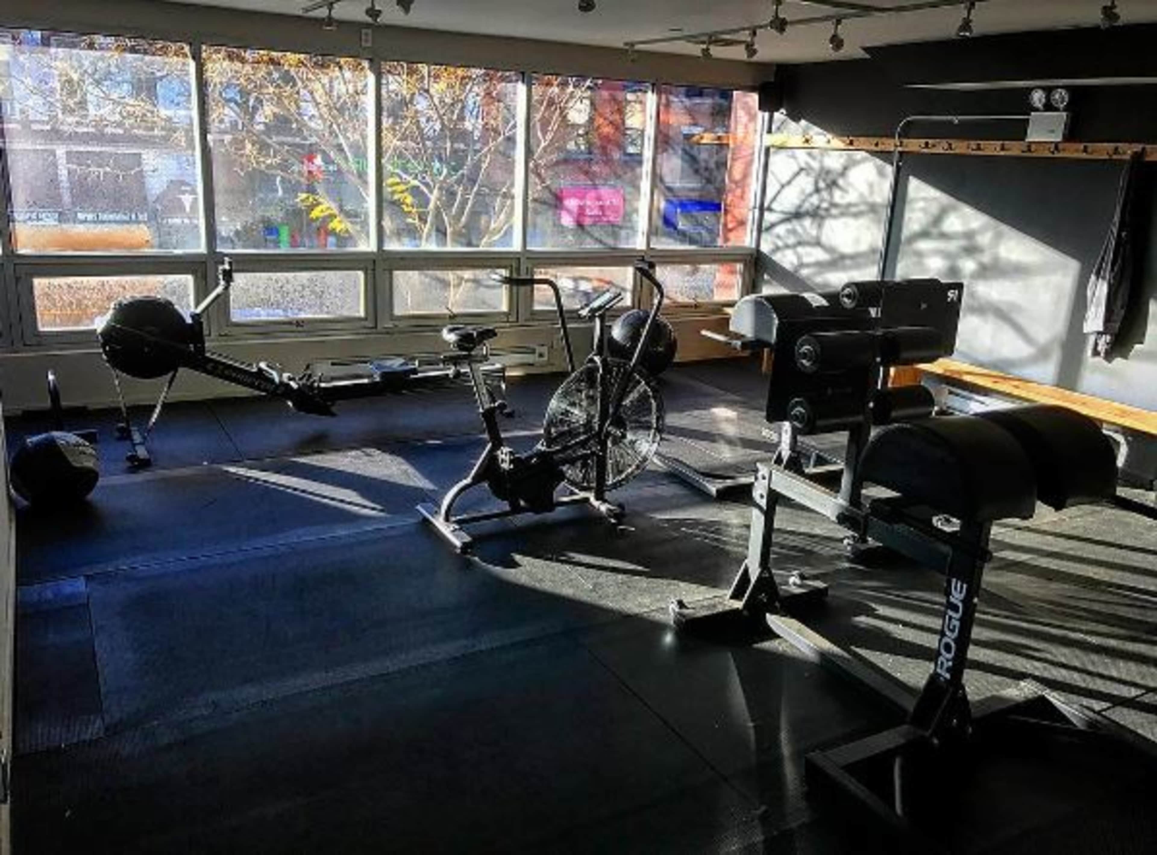 A fitness room with exercise equipment including a rowing machine, a stationary bike, and a weight bench, illuminated by sunlight through large windows.