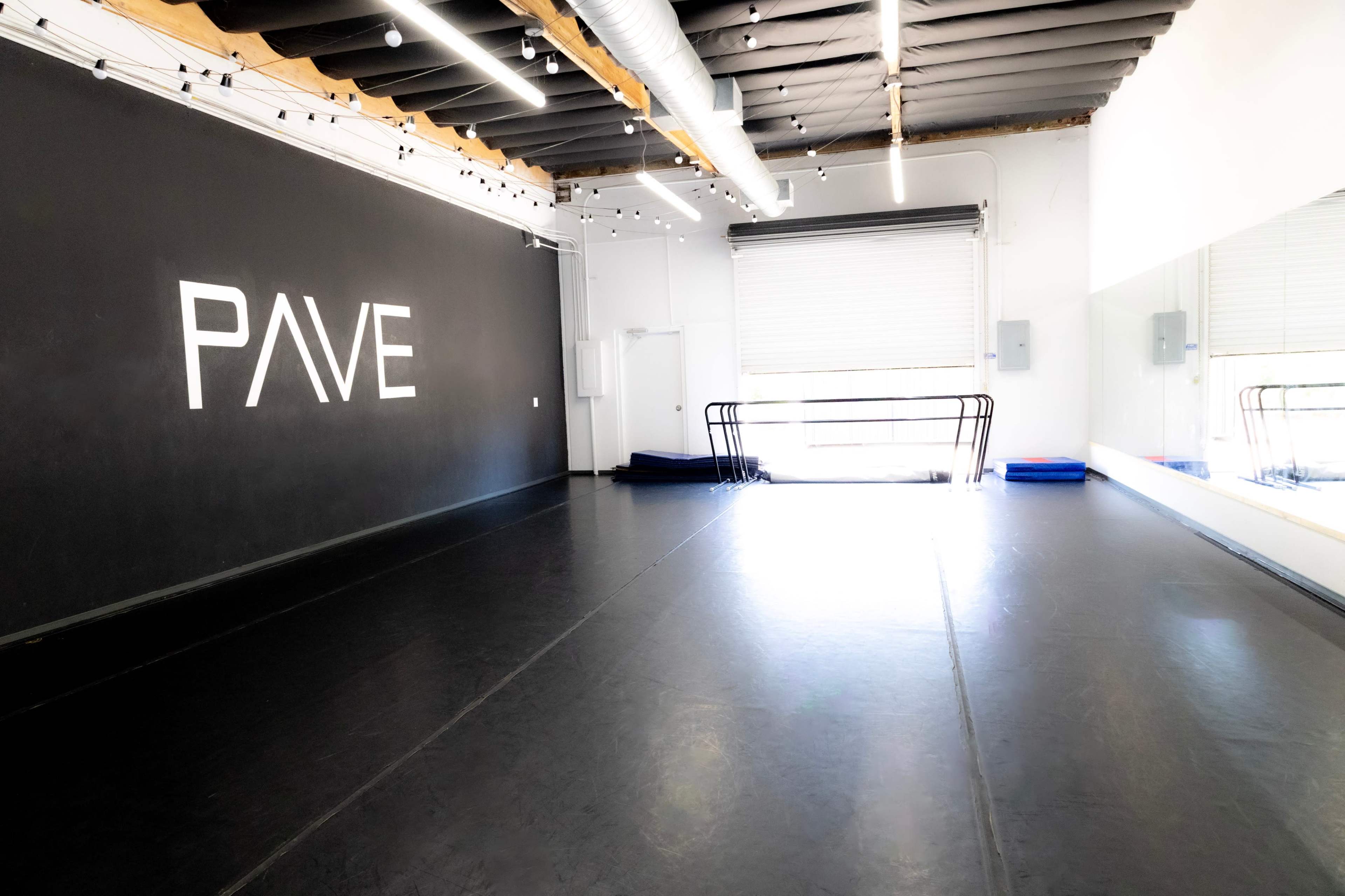 The image shows a spacious dance studio with a black wall featuring the word "PAVE," a large mirrored wall, and natural light coming through a window with a roll-up door.