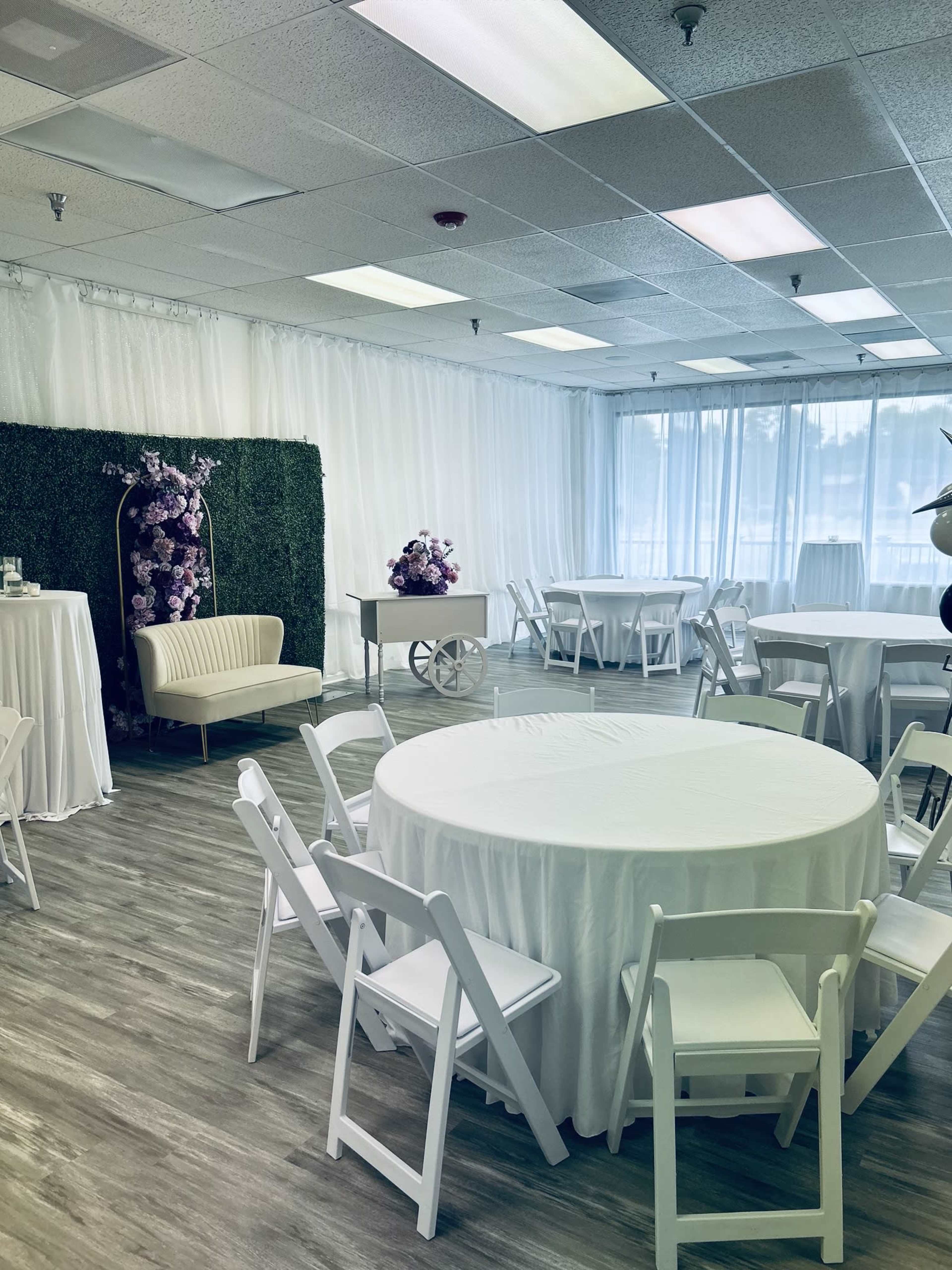 The image shows a well-decorated event space featuring round tables with white tablecloths, chairs, and a green hedge backdrop with floral arrangements.