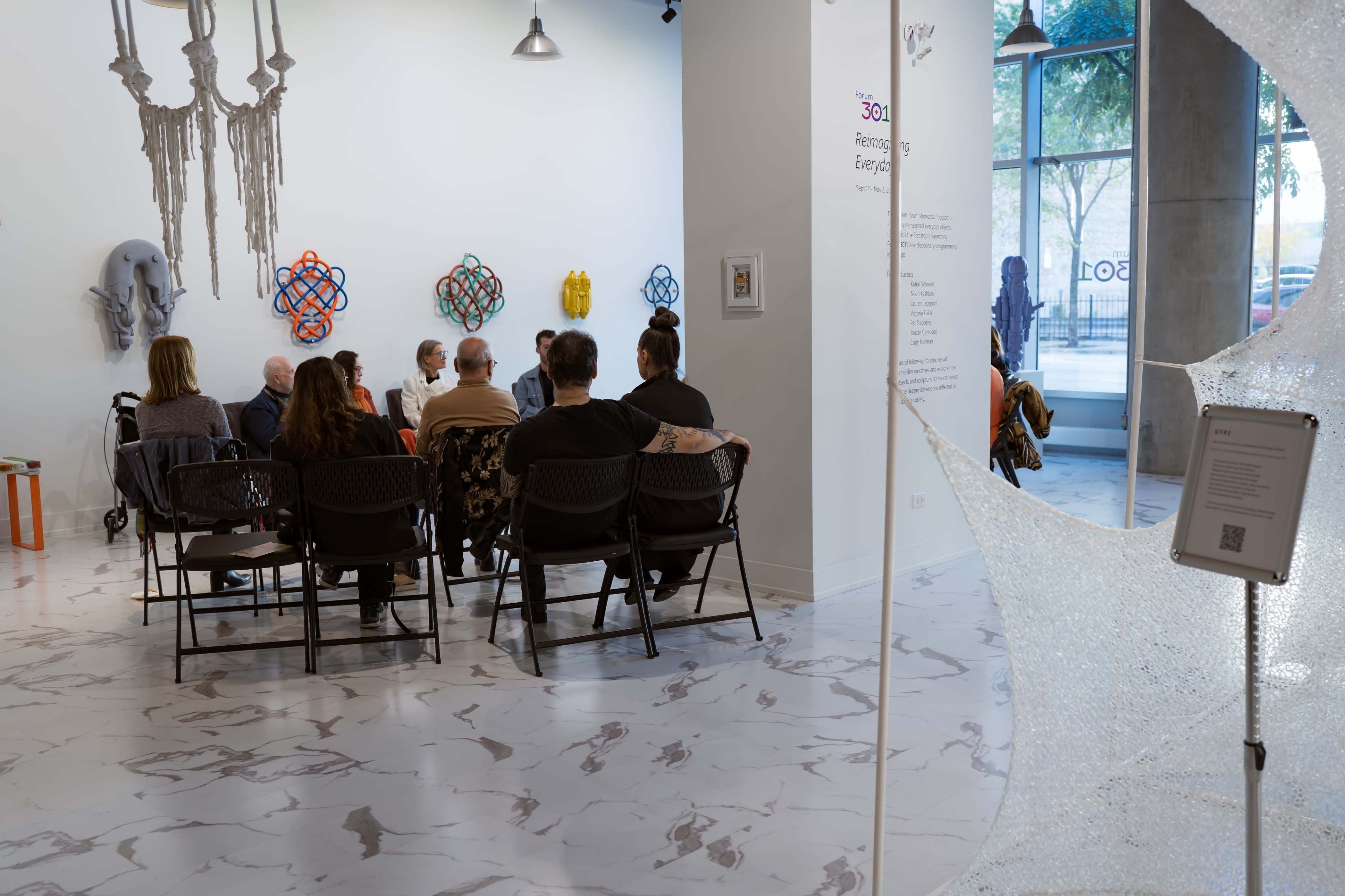 A group of people sits in chairs facing a wall adorned with various artworks in a gallery space.