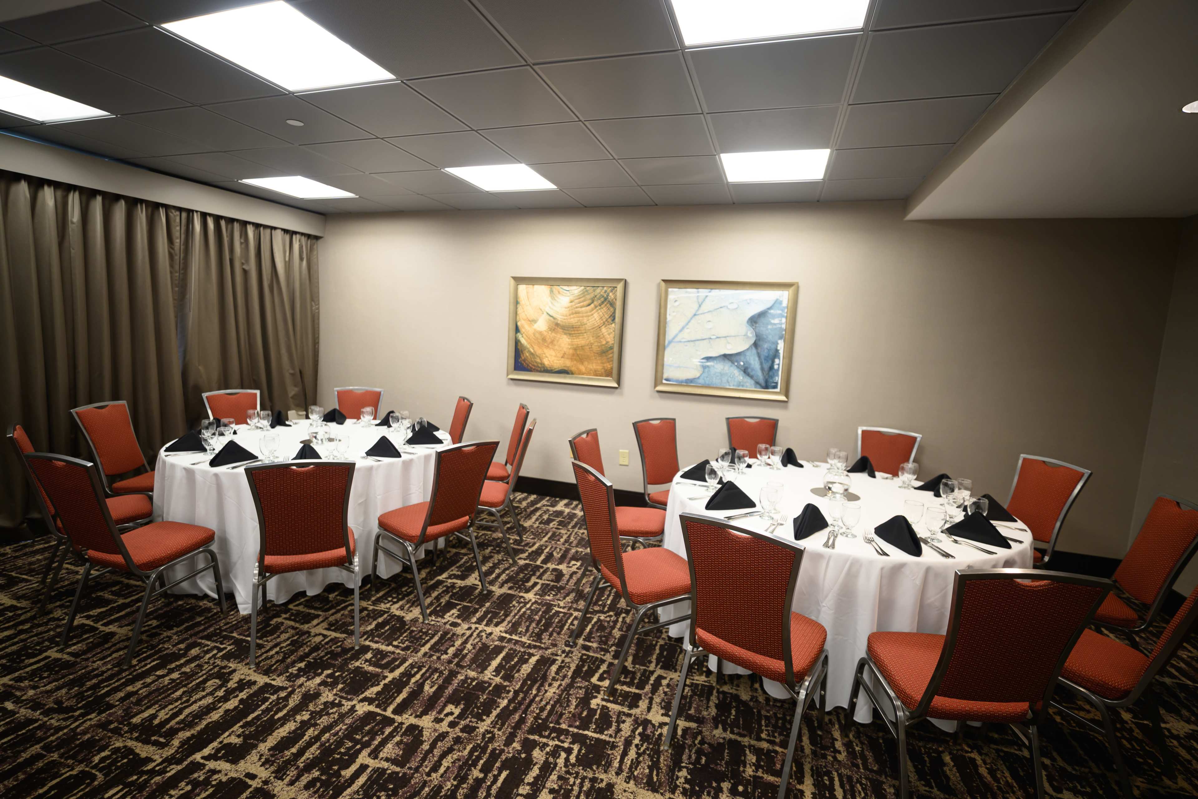 A small conference room features two round dining tables covered with white tablecloths and set with formal place settings, accompanied by red chairs and two framed artworks on the wall.