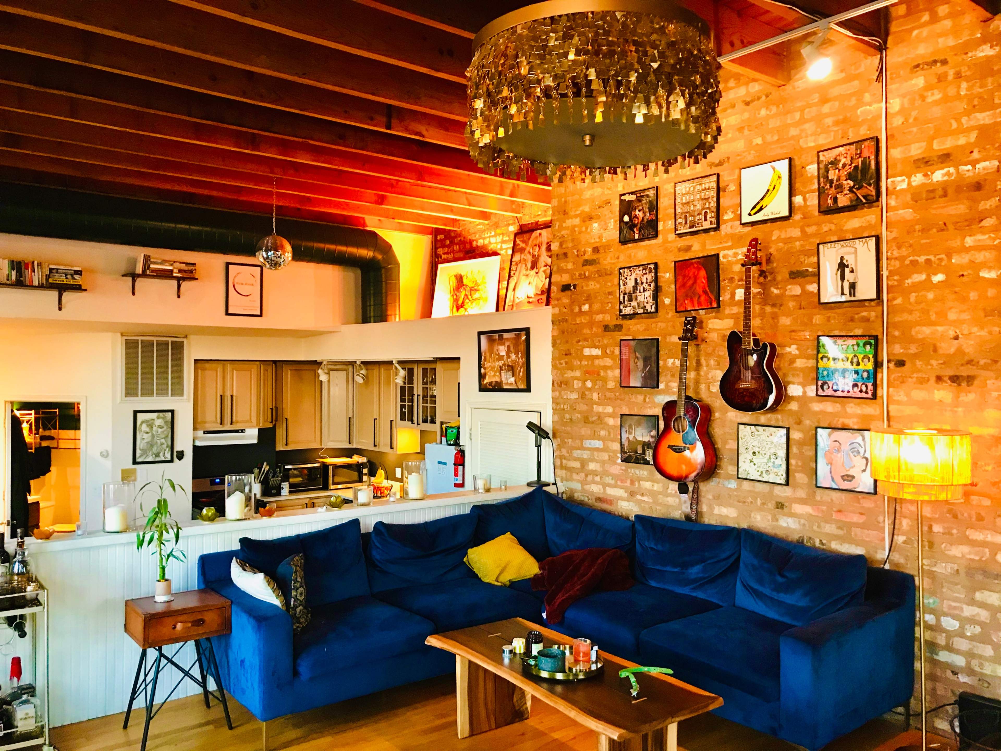 The image shows a cozy living room featuring a blue sectional sofa, a wooden coffee table, a decorative ceiling light, and various framed artworks on a brick wall.