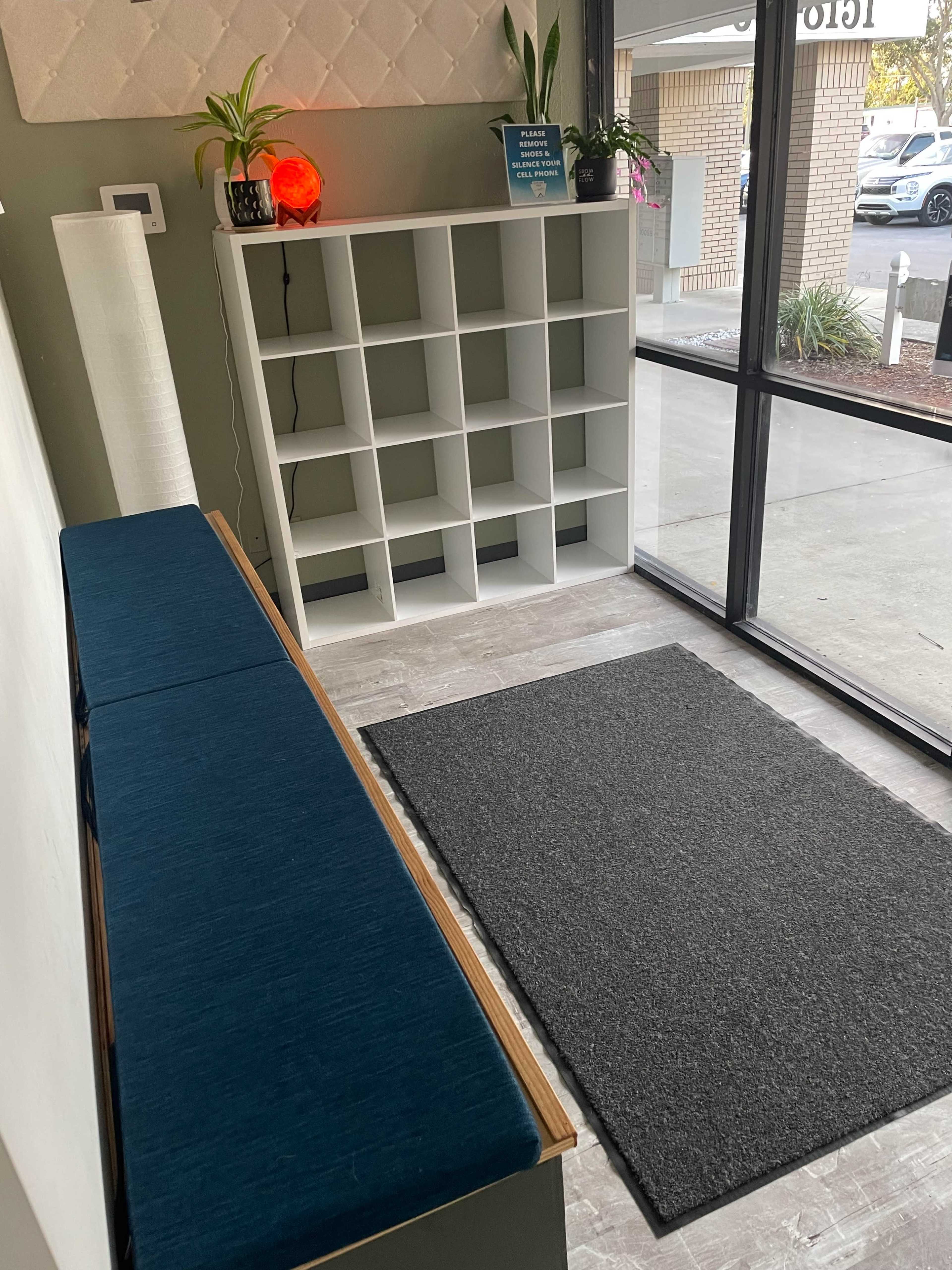 The image shows a modern entryway featuring a blue upholstered bench, a white shelving unit with multiple cubbies, a black rug, and large glass windows allowing natural light.
