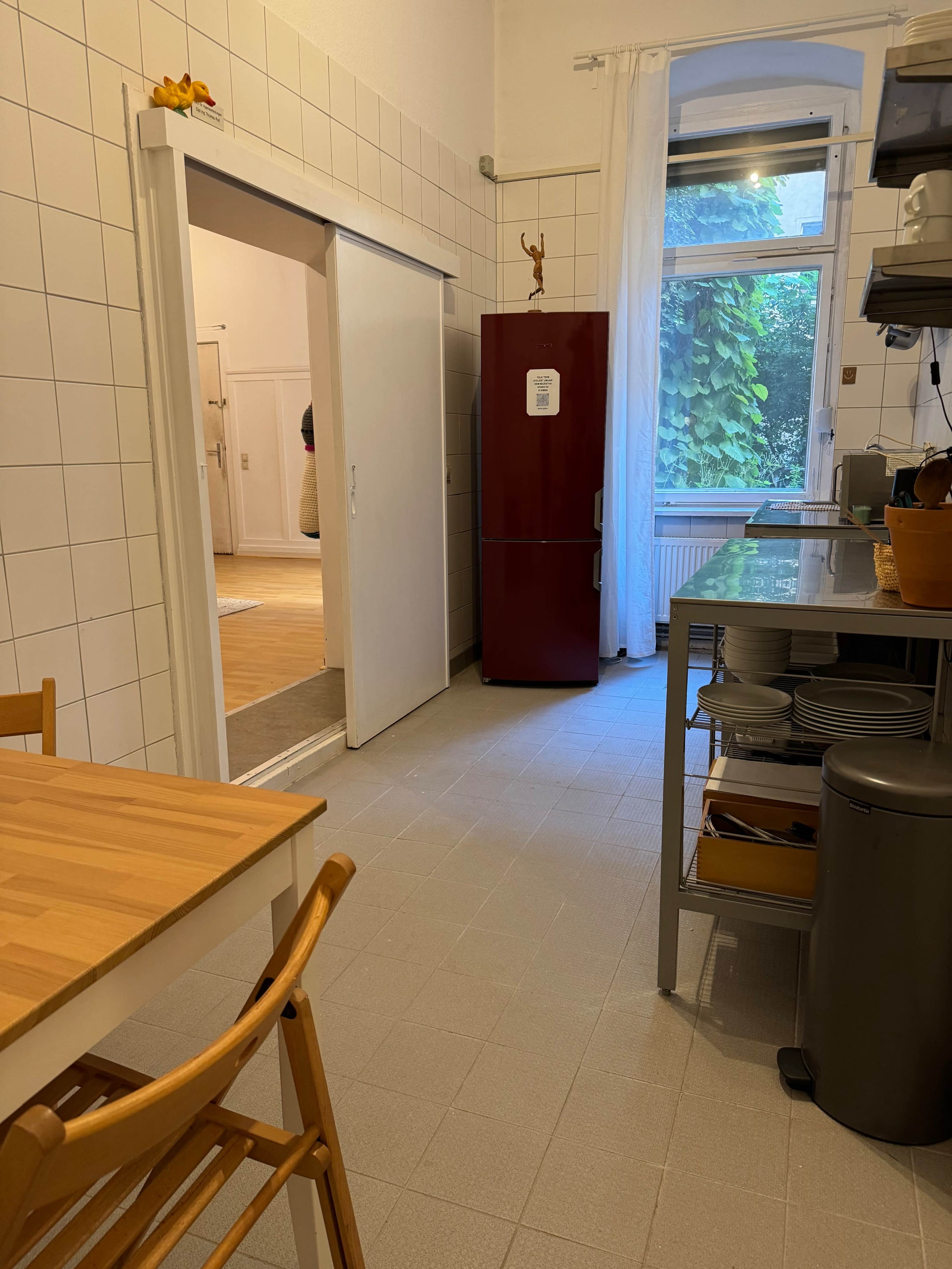 The image shows a kitchen area with a wooden dining table and chairs, a red refrigerator, and an open doorway leading to another room.