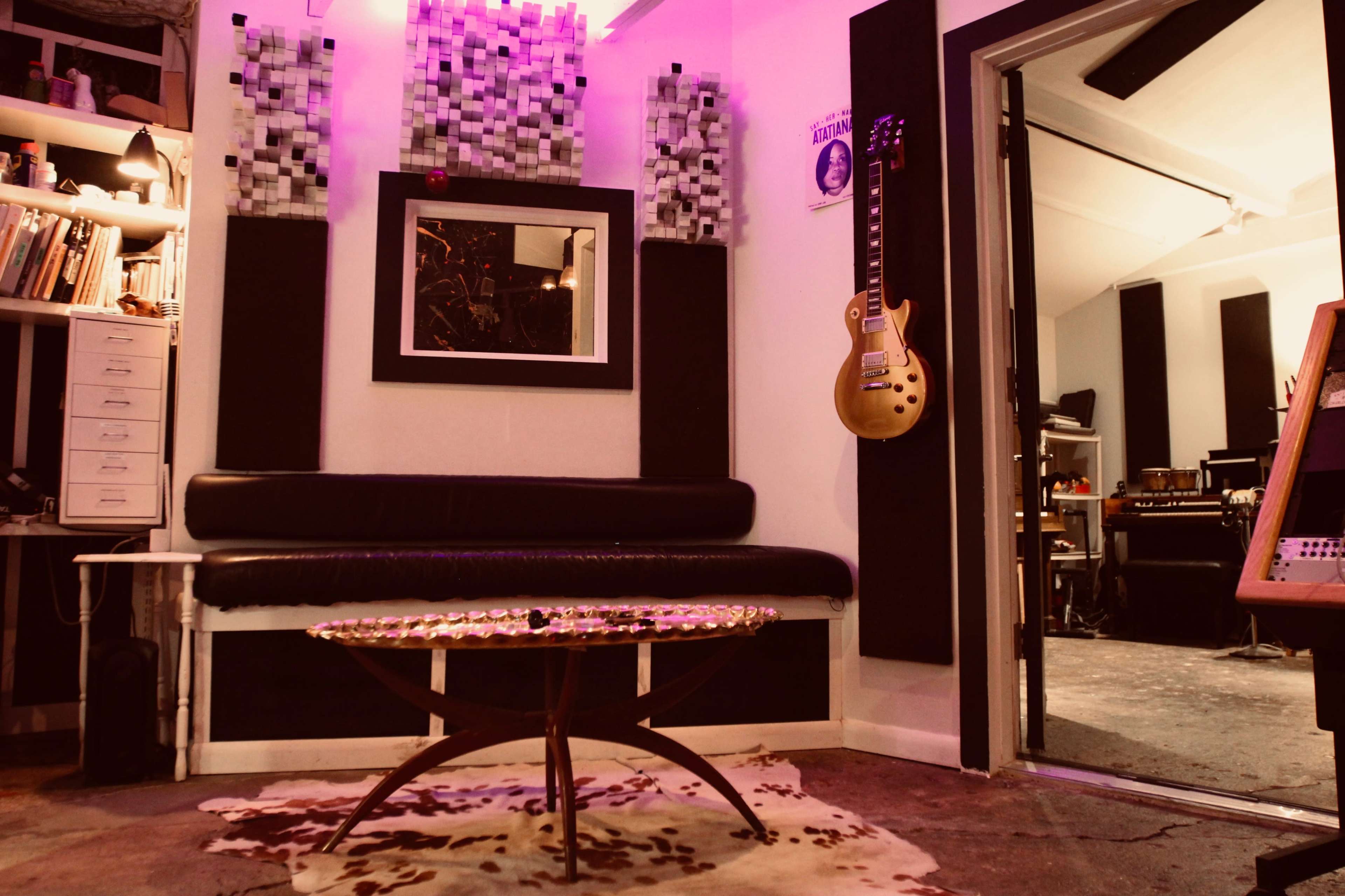The image shows a music studio with a black couch, a circular coffee table, soundproof panels on the walls, and a guitar hanging nearby.