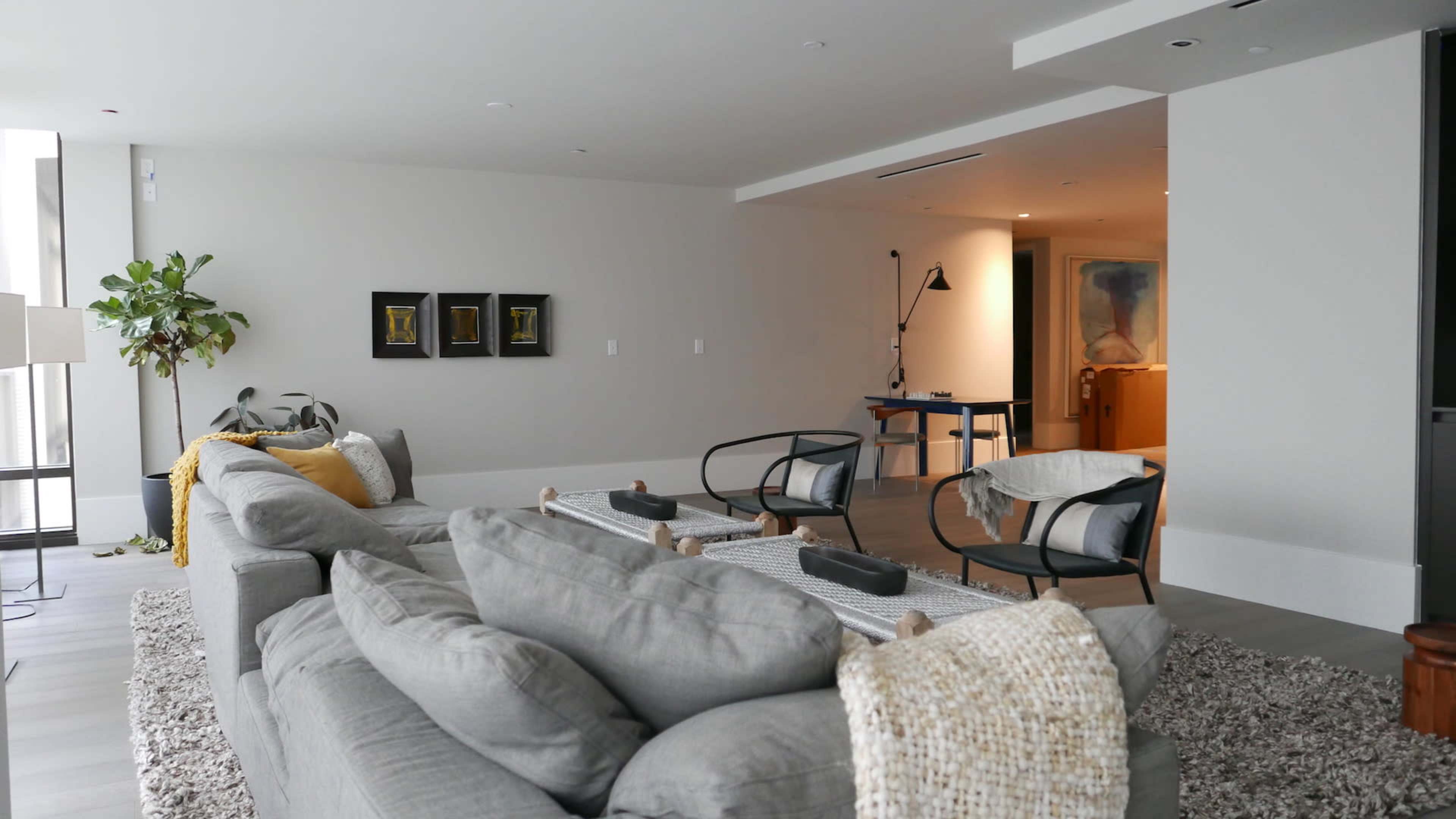 The image shows a modern living room with a gray sectional sofa, a textured rug, and minimalist decor elements, including a potted plant and artworks on the wall.