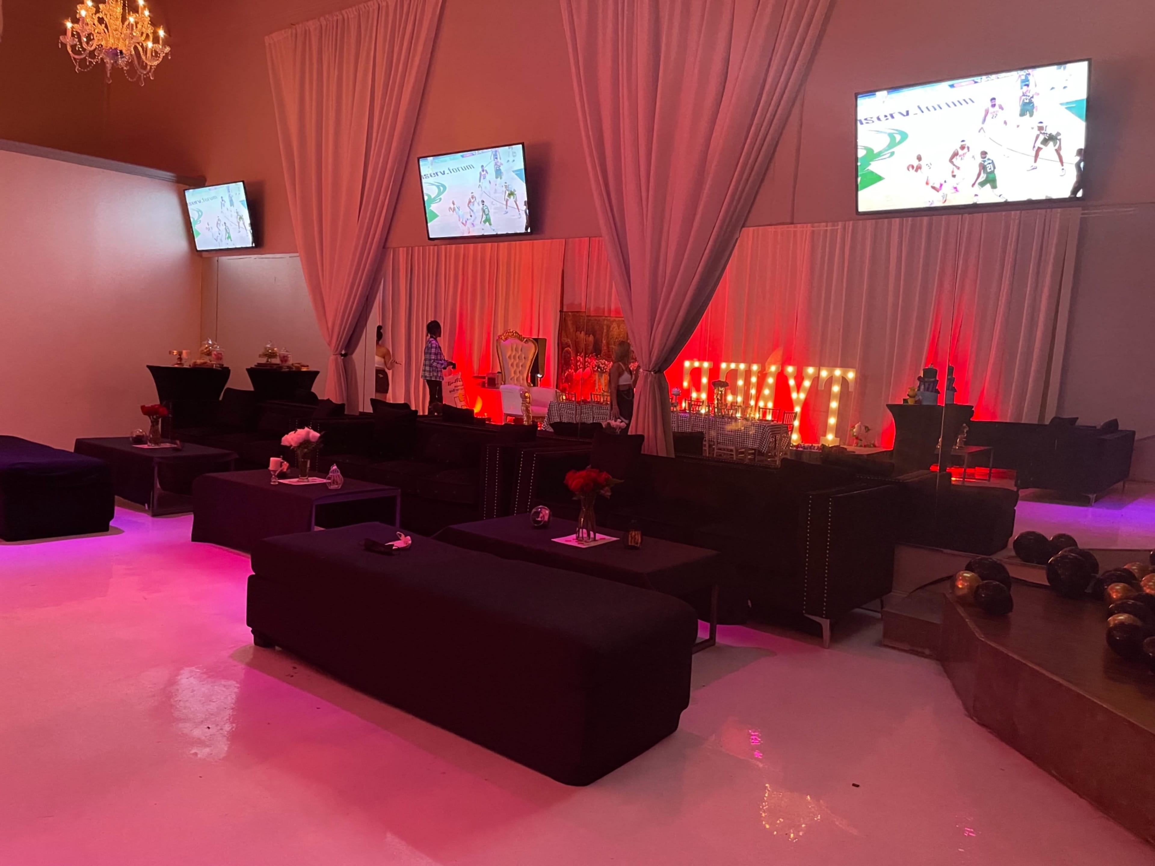 A decorated event space with black seating, illuminated red and pink lighting, and two large screens displaying a basketball game.