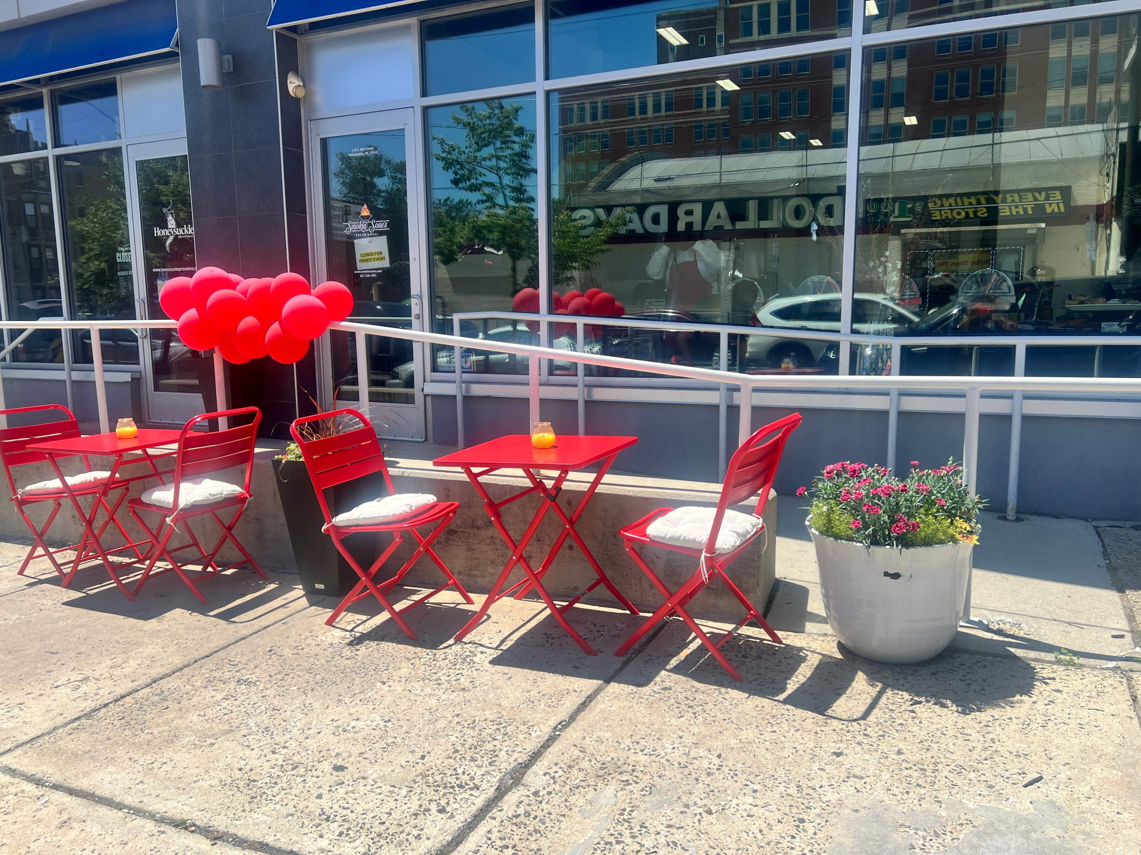 A row of red tables and chairs is set up outside a storefront, accompanied by a potted plant and red balloons.
