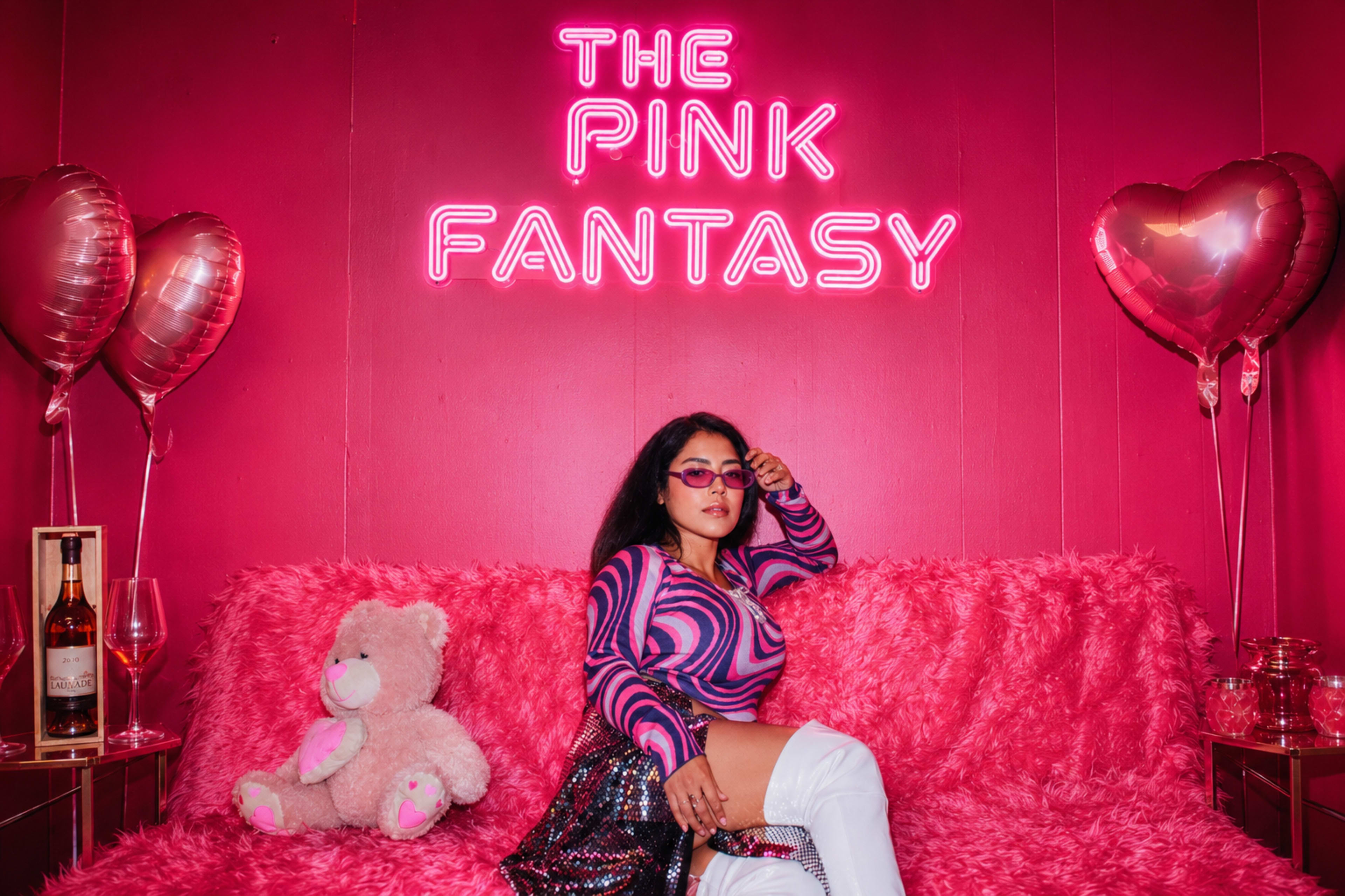 A woman in a colorful striped sweater and thigh-high white boots sits on a pink faux-fur sofa, posing next to a teddy bear and illuminated by a neon sign that reads "THE PINK FANTASY."