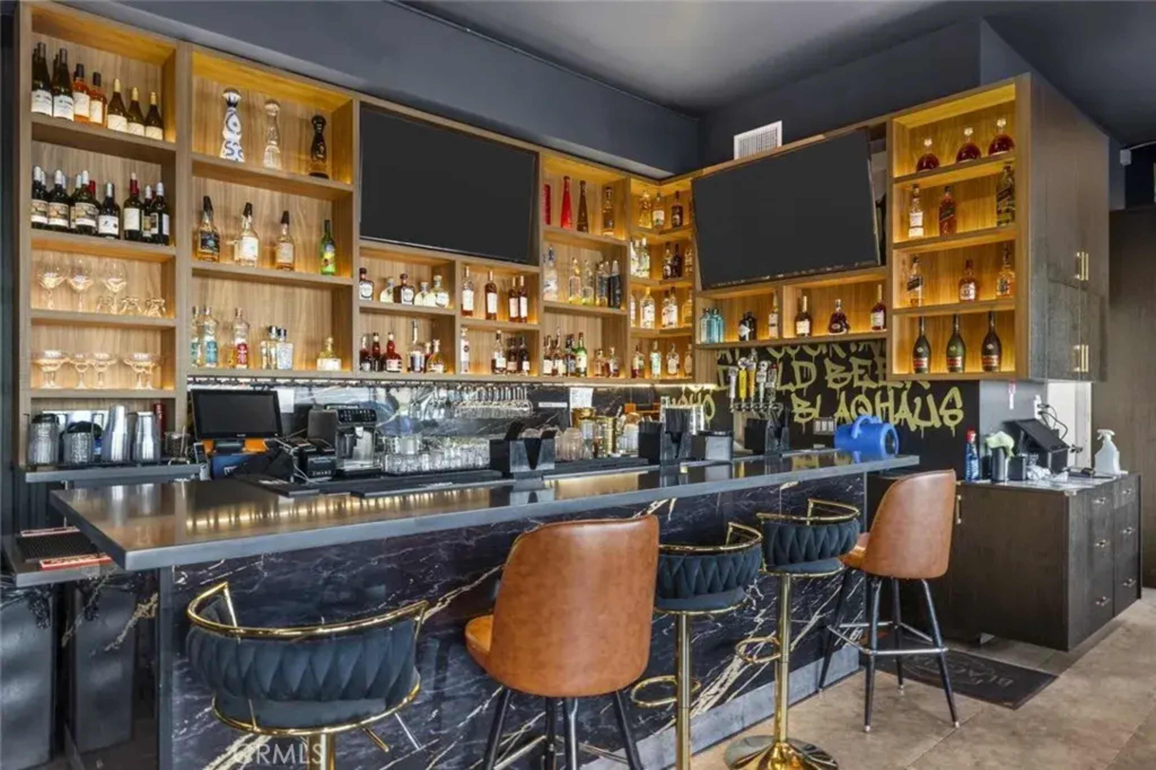 The image shows a modern bar setup featuring a marble counter, high-backed chairs, and shelves stocked with various alcoholic beverages, with multiple screens mounted on the wall.