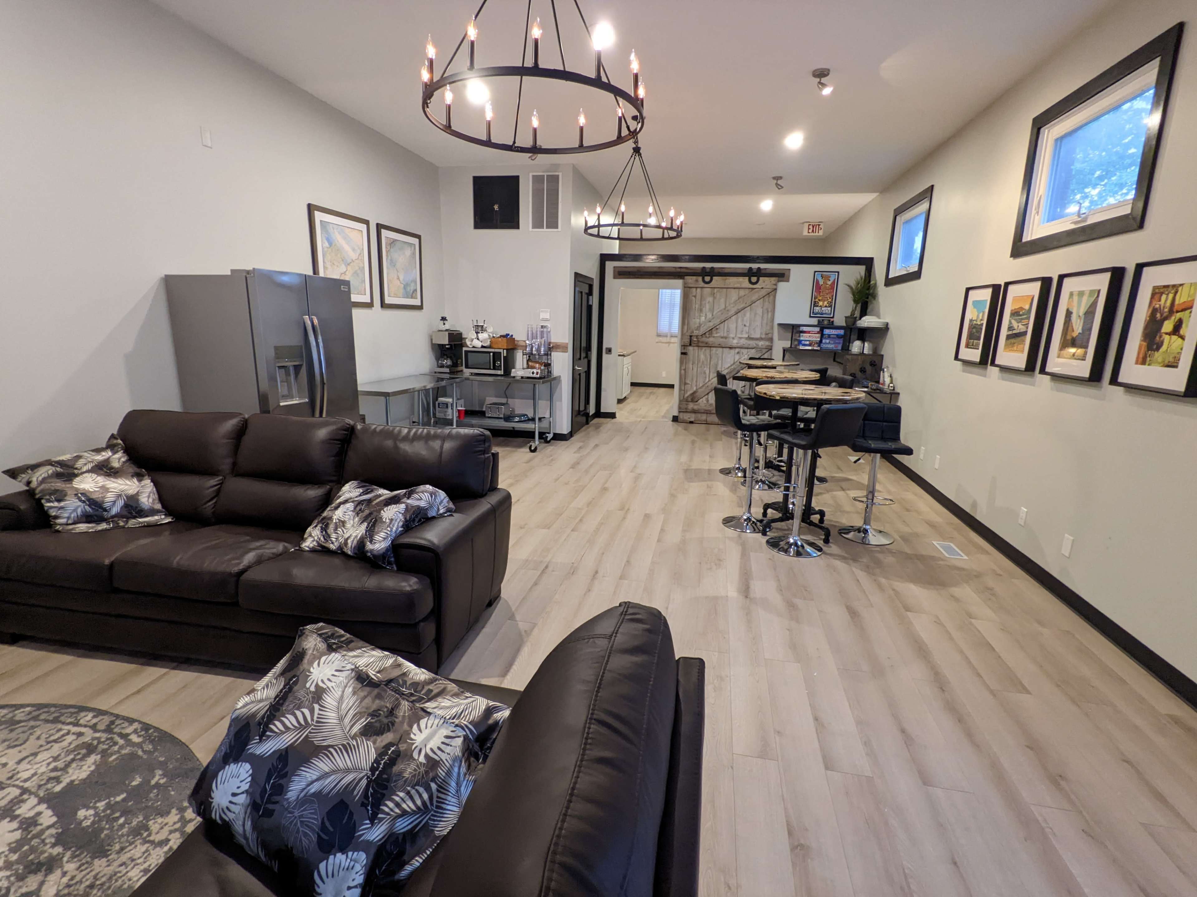 A modern, open living space featuring two black couches, a dining area with bar stools, and a kitchenette with stainless steel appliances.