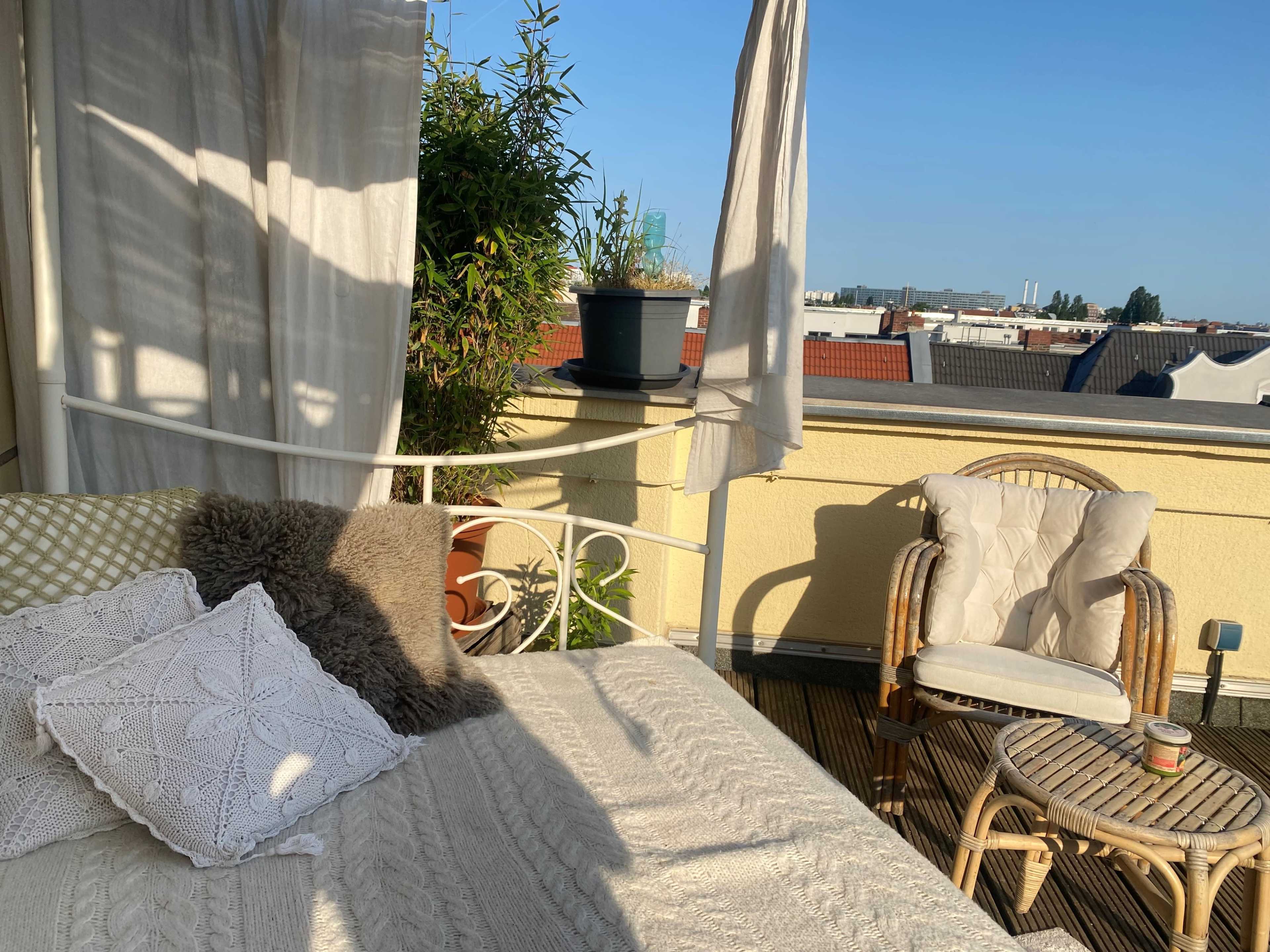 A sunlit balcony features a white metal bed with pillows, a wicker chair, and potted plants against a clear blue sky.