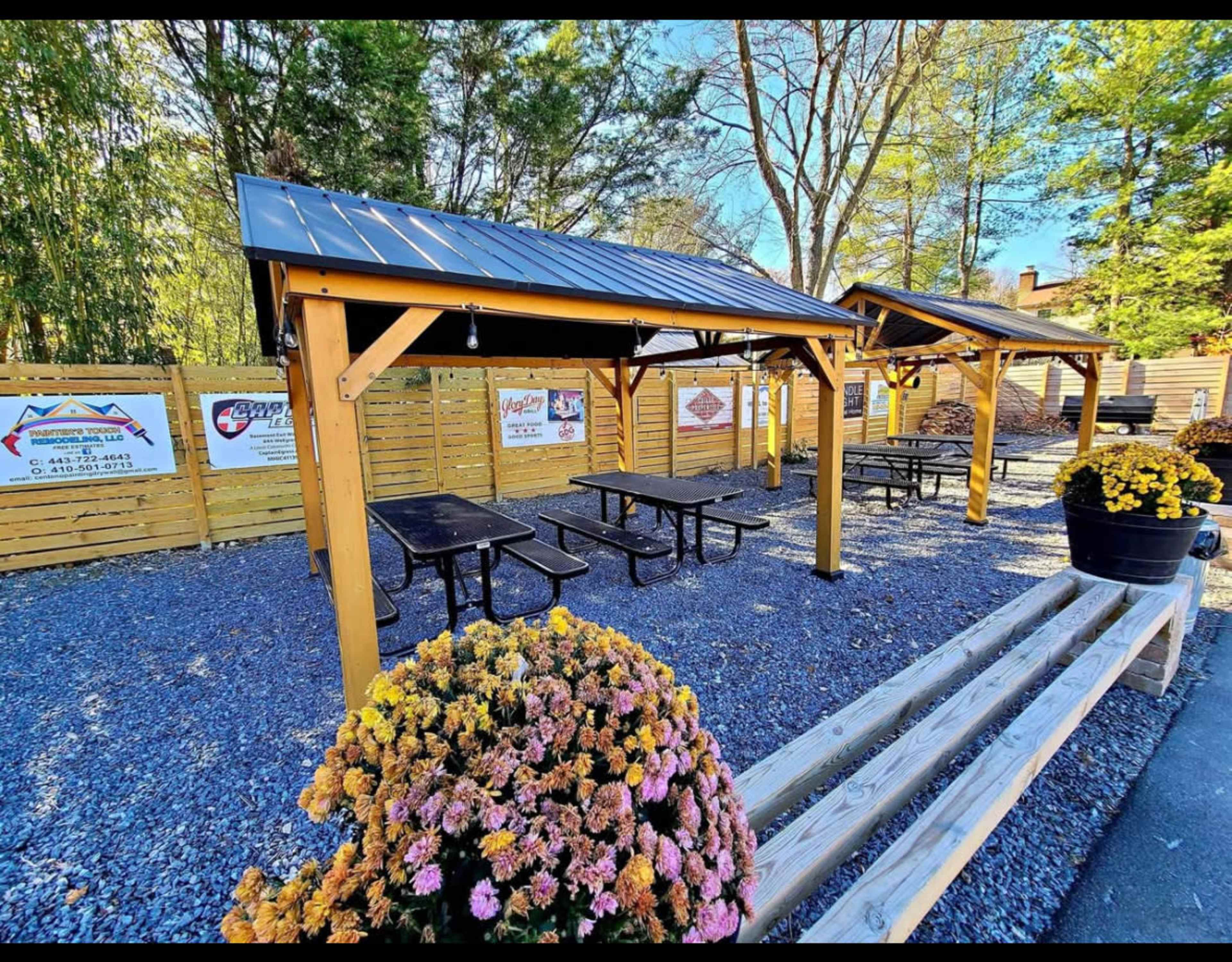 The image shows an outdoor area with multiple picnic tables under wooden pavilions, surrounded by a gravel surface and fenced by wooden panels.