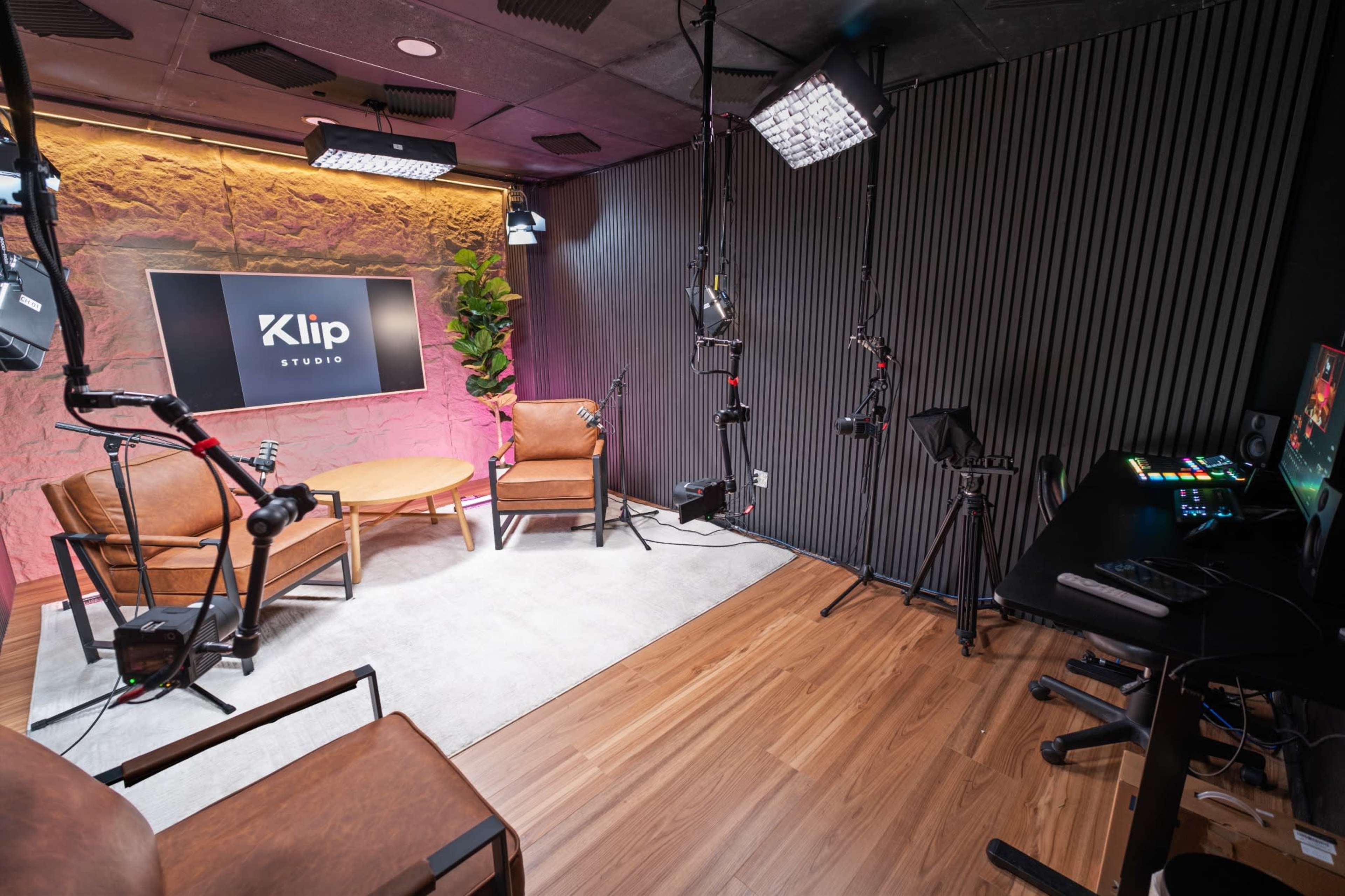 The image shows a modern studio setup featuring two brown armchairs, a round coffee table, various lighting equipment, and a control desk with monitors, all arranged for video production.