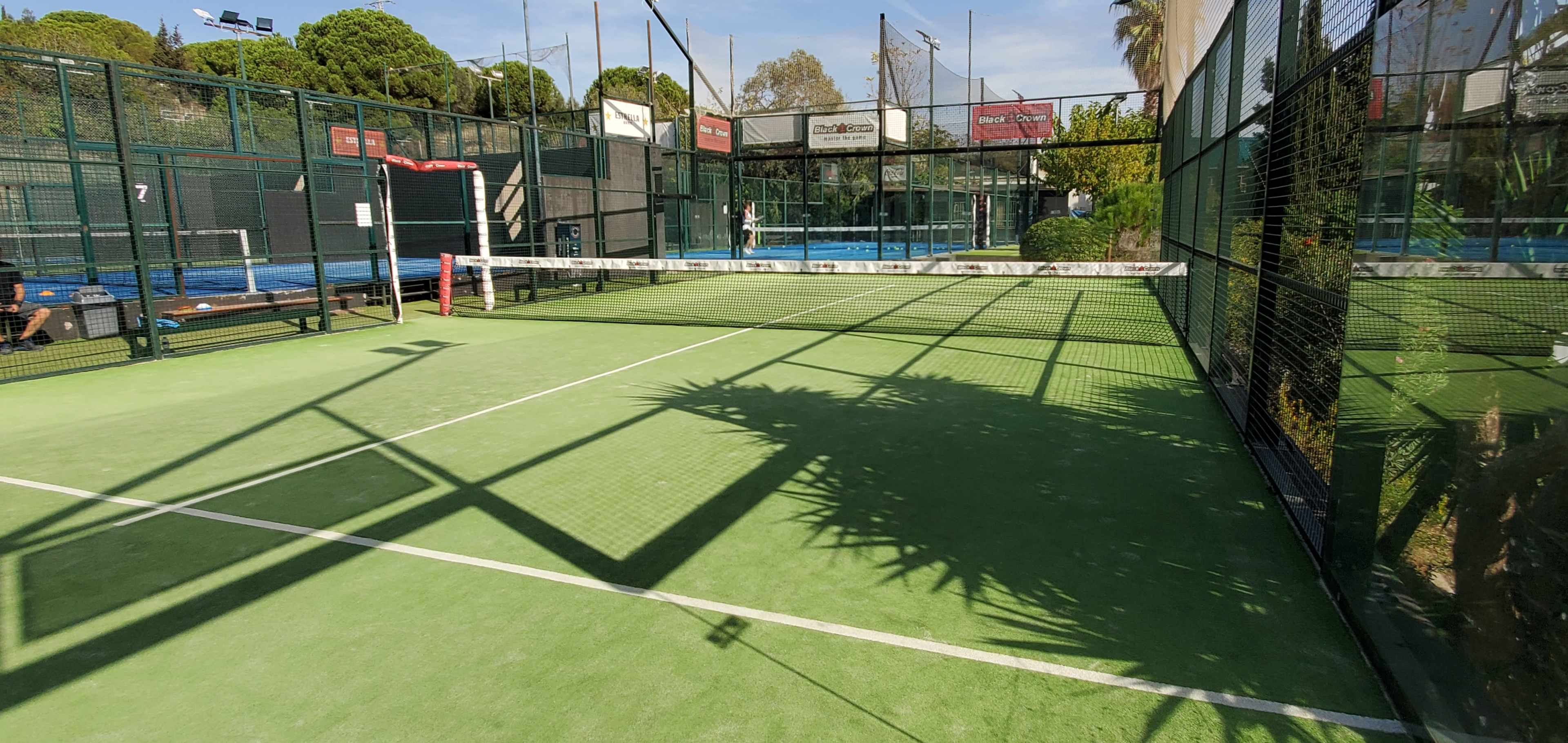 A padel tennis court with green flooring, a net dividing the playing area, and shadows cast by surrounding structures.