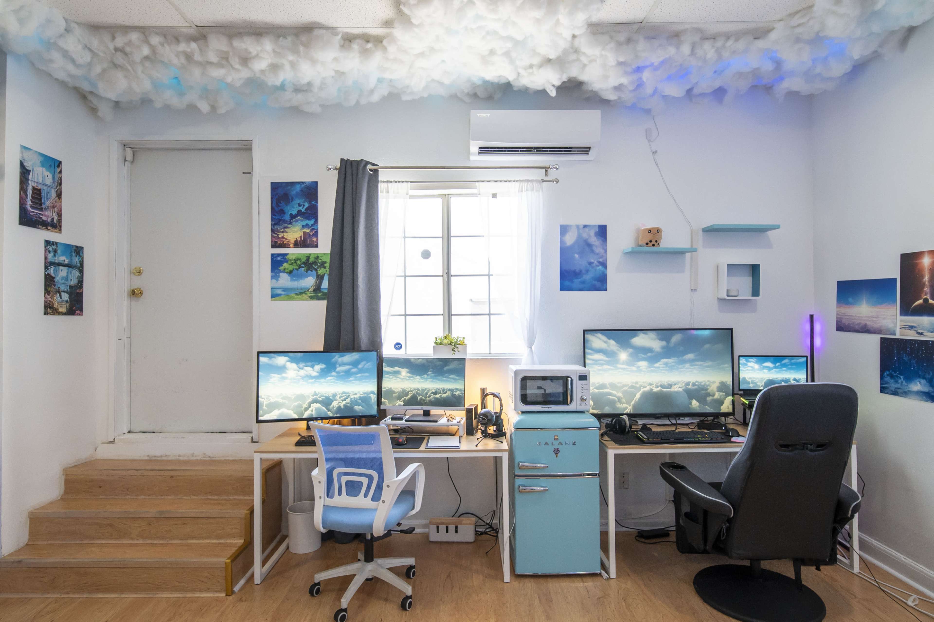 A bright office space features two desks with multiple computer monitors, an air conditioning unit, and cloud-themed decor on the ceiling.