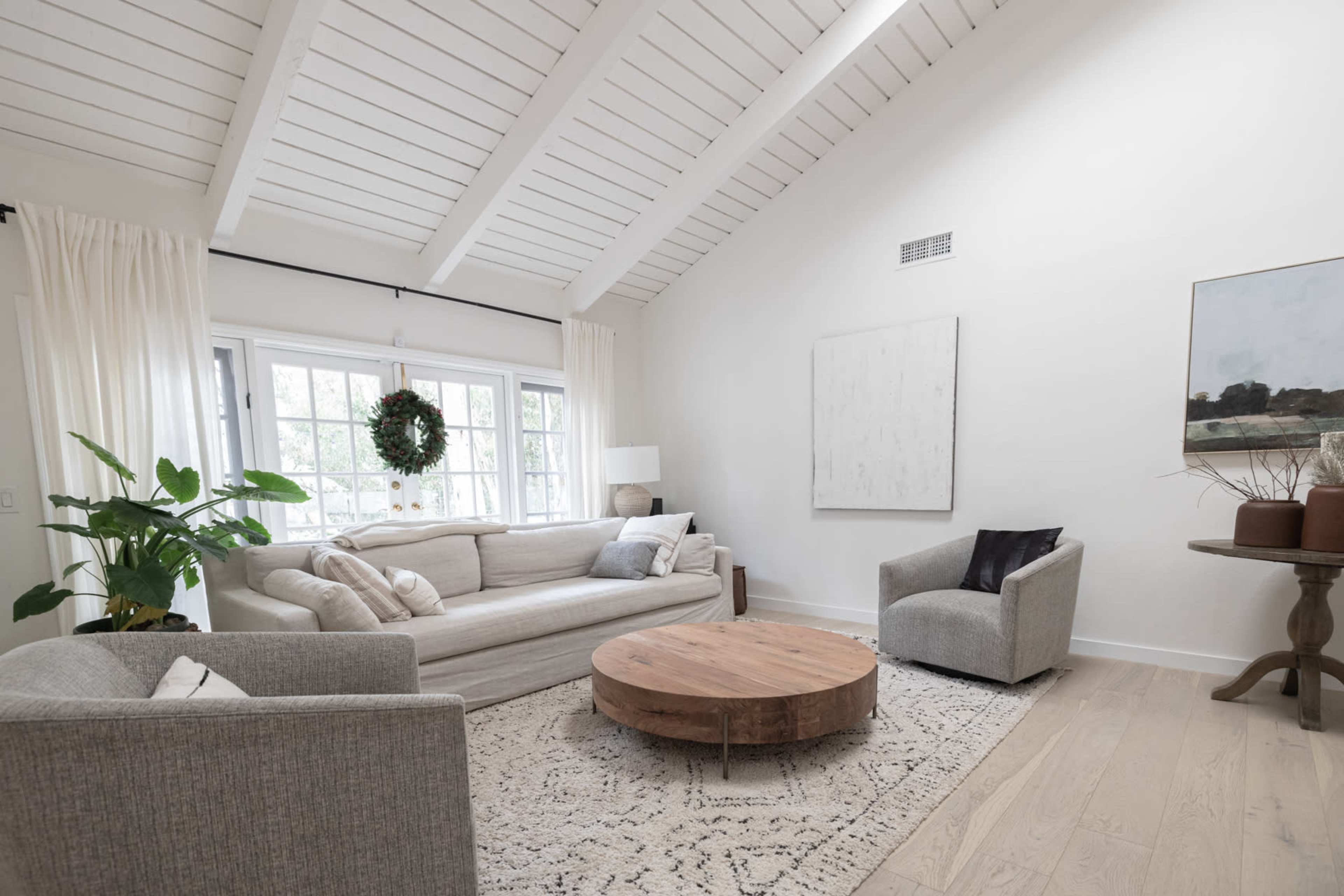 A bright living room featuring a light-colored sofa, two armchairs, a round wooden coffee table, and large windows with natural light.