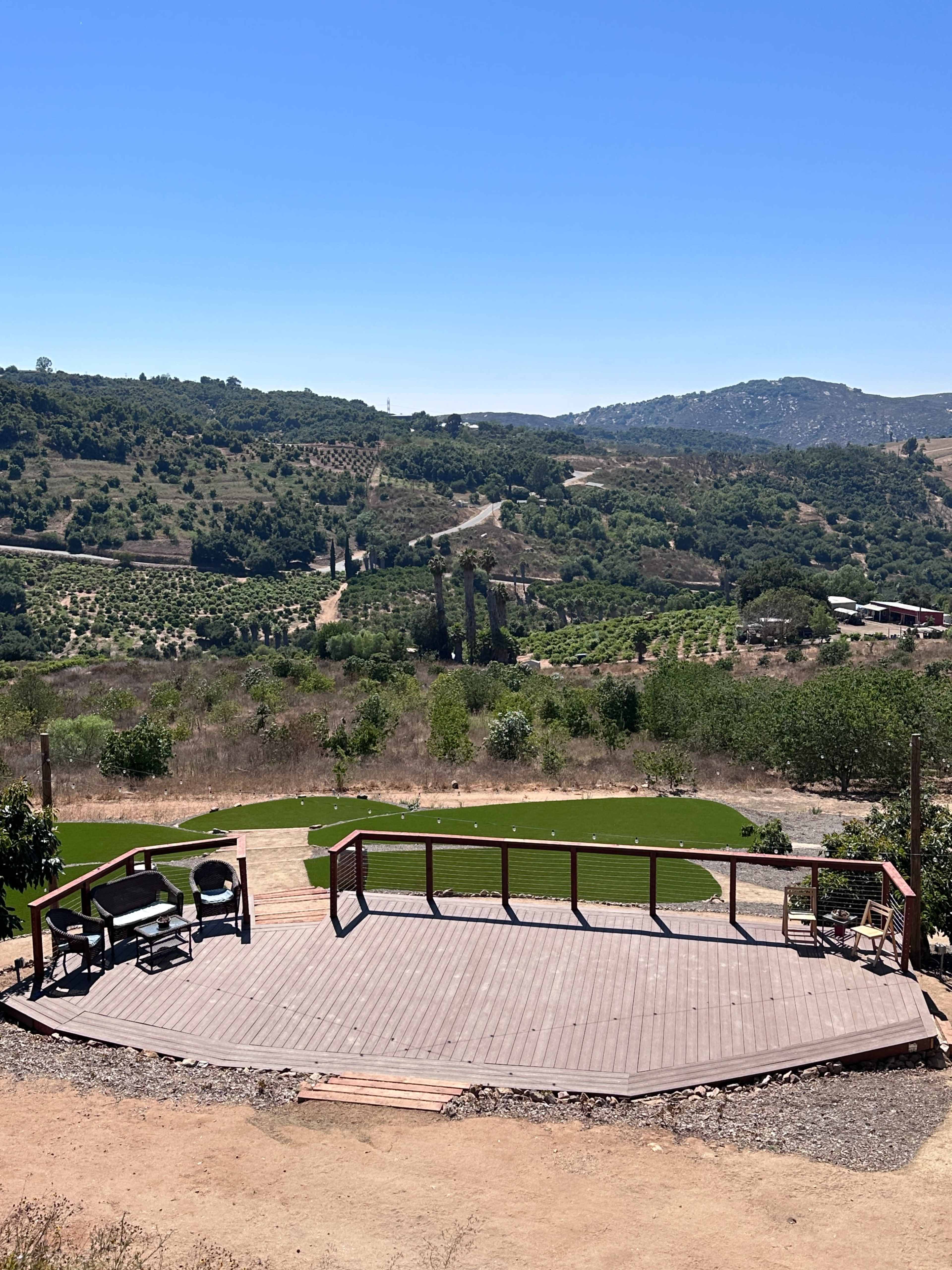 A wooden deck overlooks a green valley with rows of trees and a winding road in the distance.
