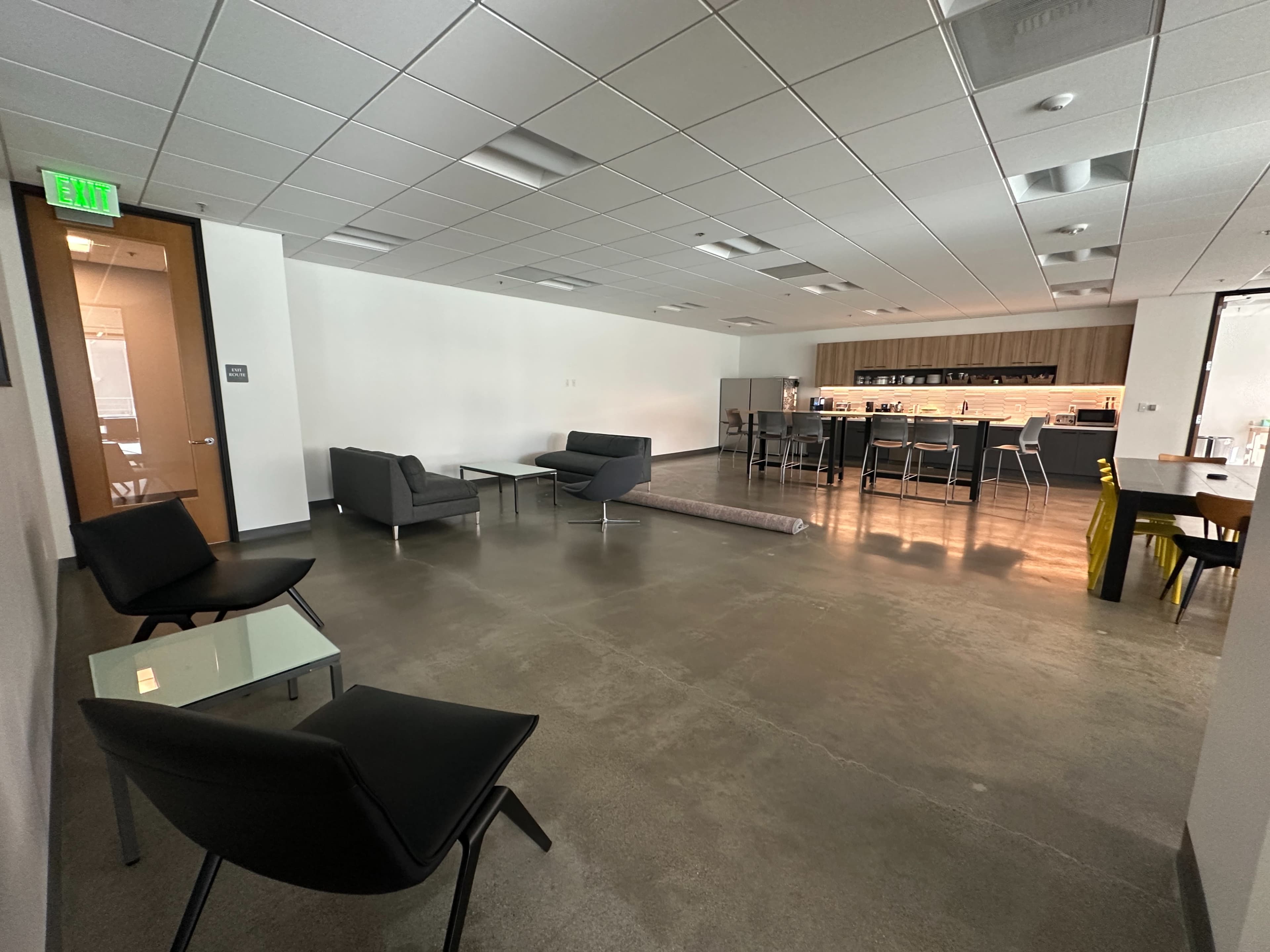 The image shows a modern office space featuring seating arrangements with gray couches and black chairs, a counter area with bar stools, and a polished concrete floor.