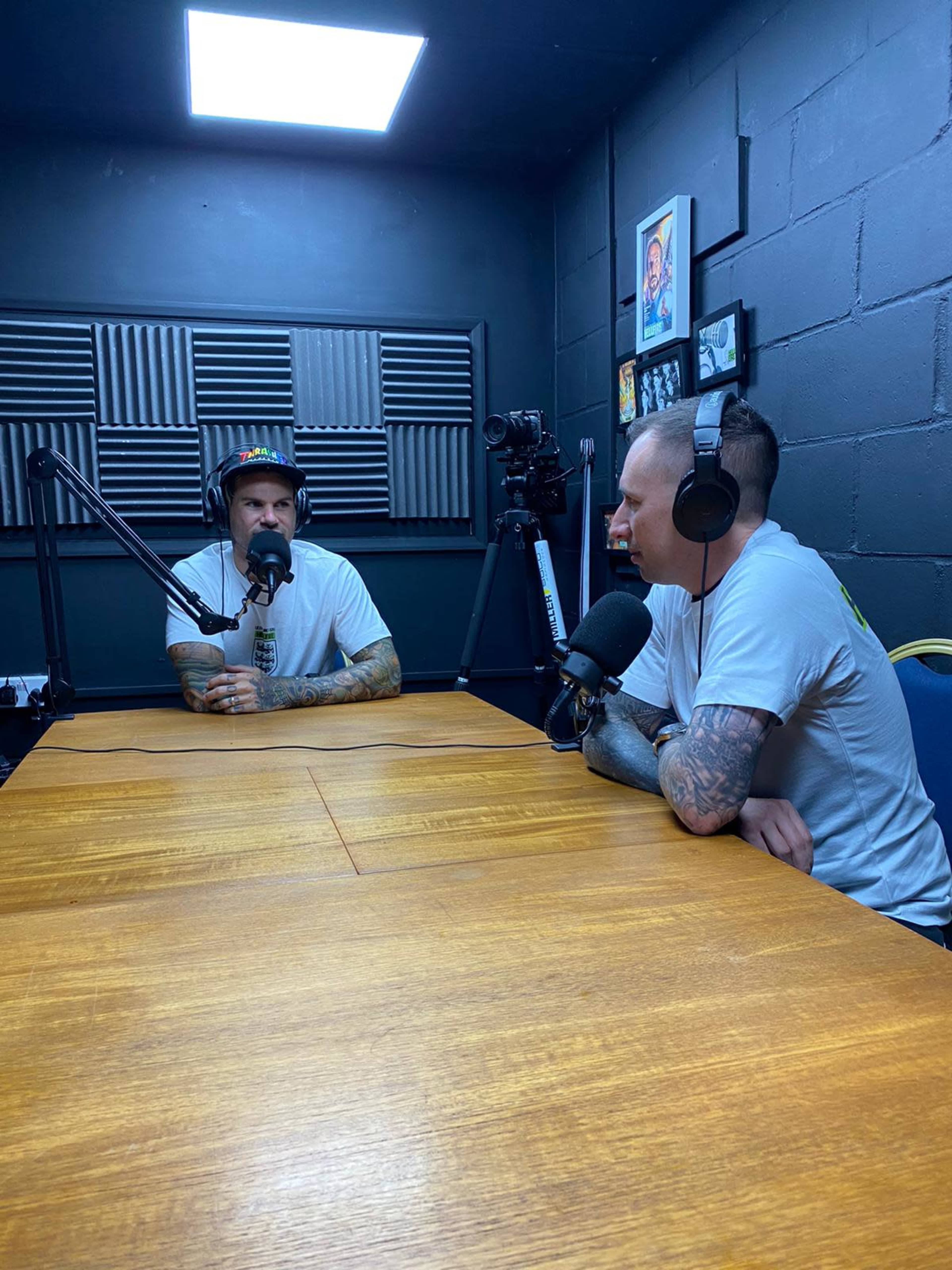 Two men with tattoos are sitting at a wooden table in a recording studio, wearing headphones and speaking into microphones.